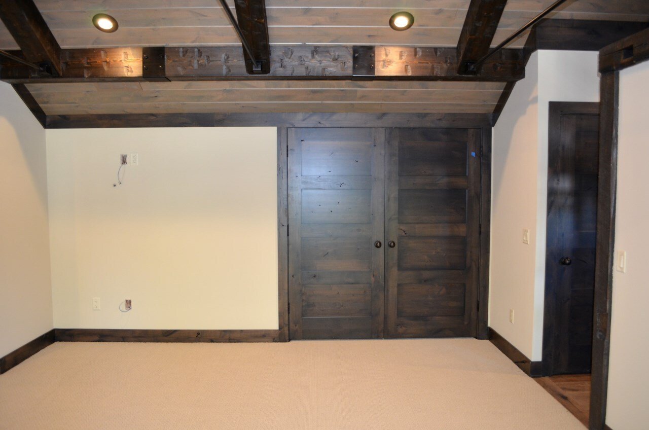 Empty room with beige carpet, white walls, dark wooden sliding closet door, and exposed ceiling beams with two ceiling lights