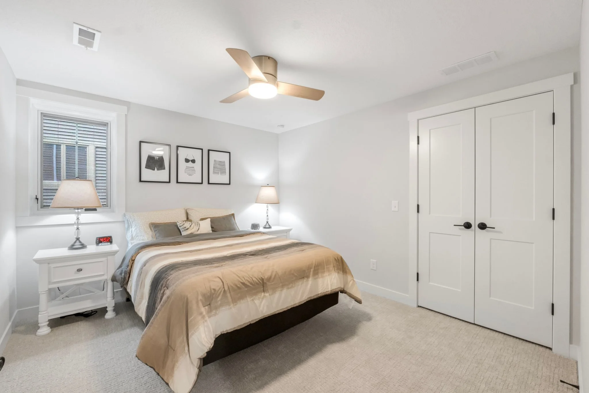A bedroom with a white bed, nightstands, two lamps, a ceiling fan, and wall art with black frames.