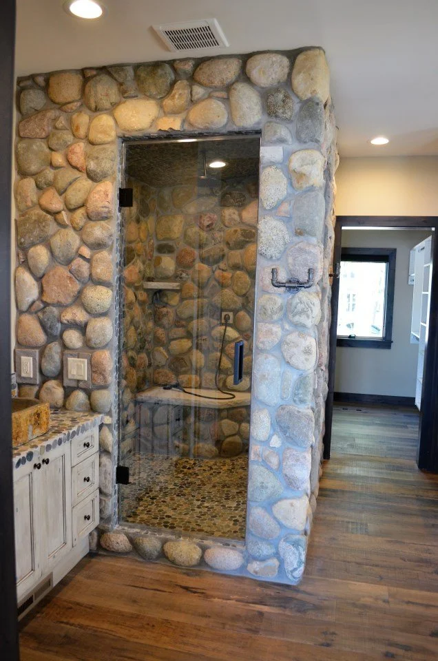 Interior of a house with a walk-in shower enclosed by glass, surrounded by stone walls, with a wooden floor and a nearby white cabinet, a hallway leading to another room and windows in the background.