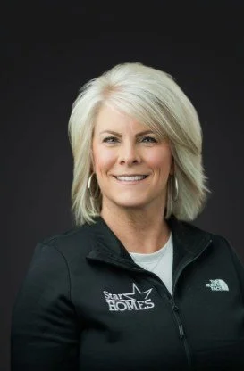 A woman with blonde hair smiling, wearing a black jacket with a logo that says 'Star Homes' and North Face branding, against a black background.