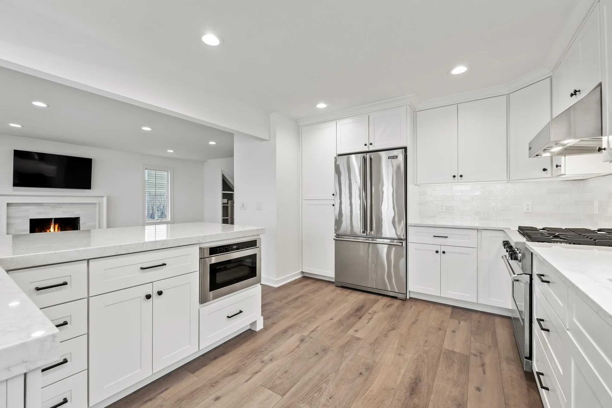 Modern white kitchen with stainless steel appliances, wooden flooring, and white cabinets. Visible are a refrigerator, oven, stovetop, and microwave, with a white backsplash and recessed lighting.