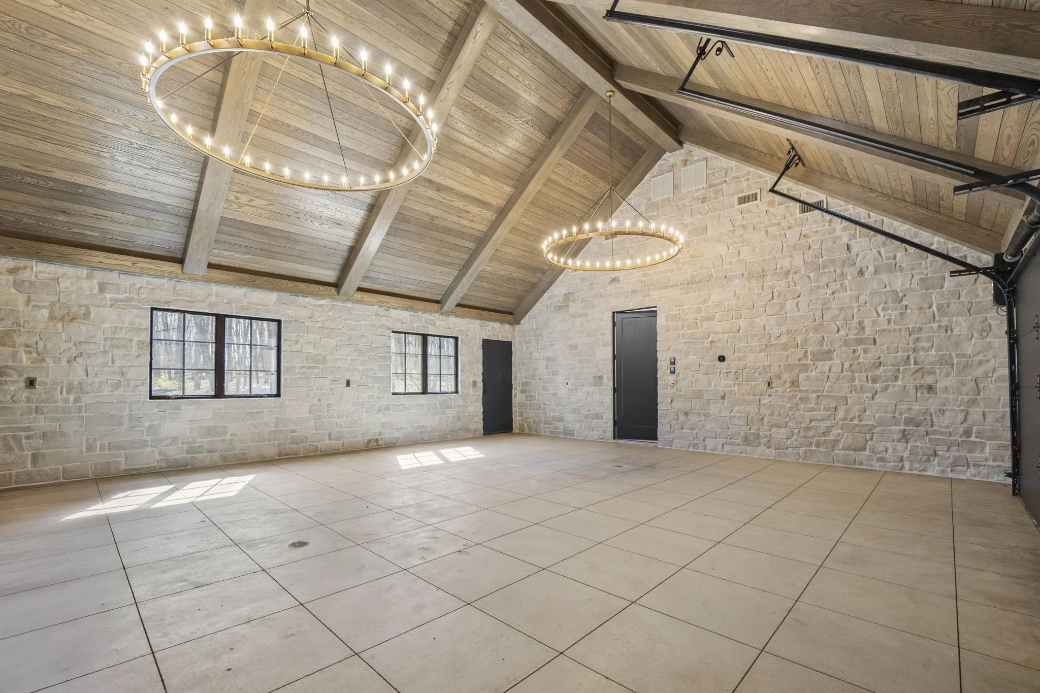 Empty room with stone walls, wooden ceiling with exposed beams, two circular chandeliers with candles, tiled floor, and black doors and windows.