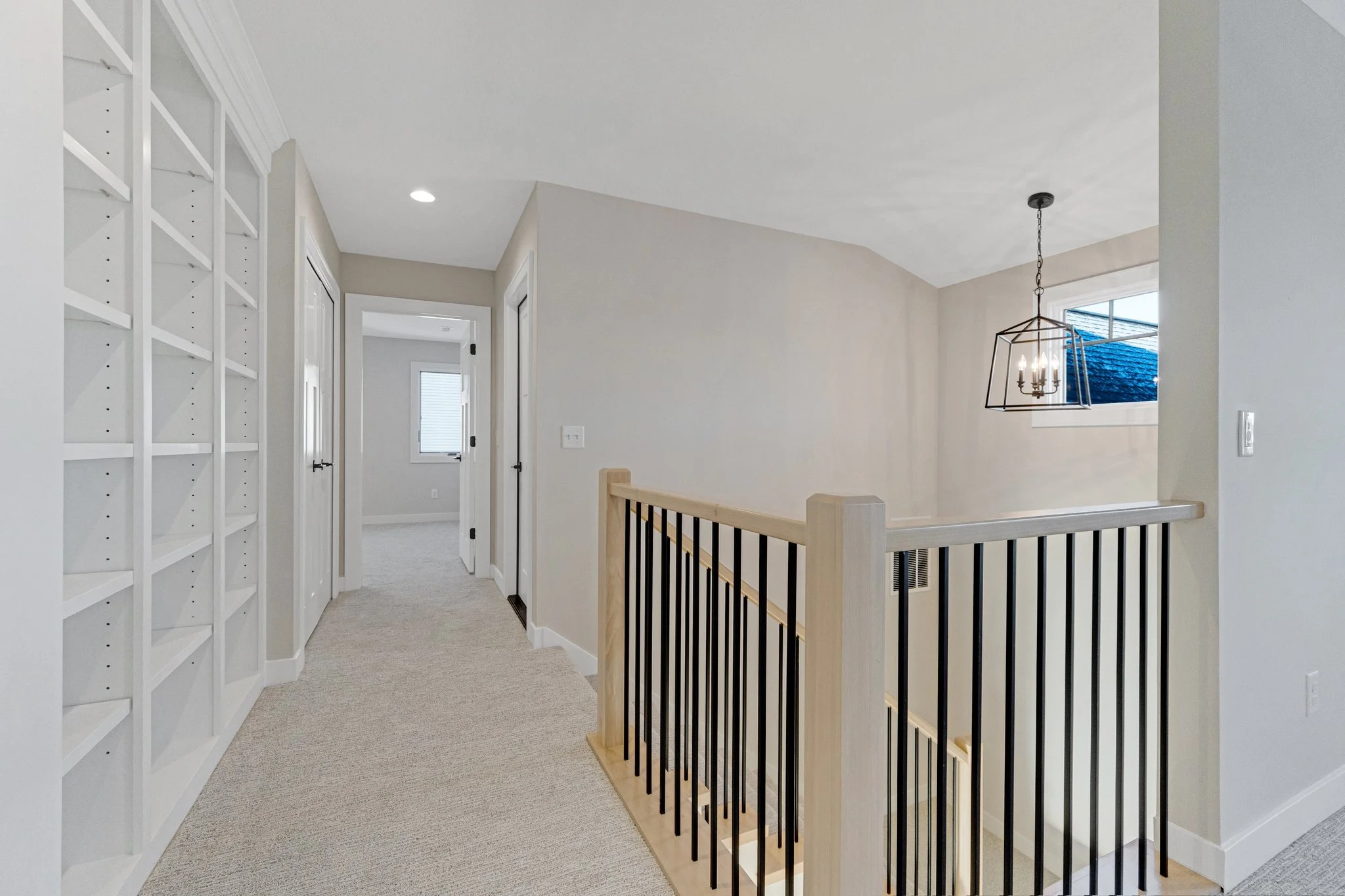 Interior of a modern house upstairs hallway with a white bookshelf, beige carpet, and a staircase with black metal and light wood railing. There are multiple doors leading to bedrooms and a window with a chandelier hanging above the staircase.