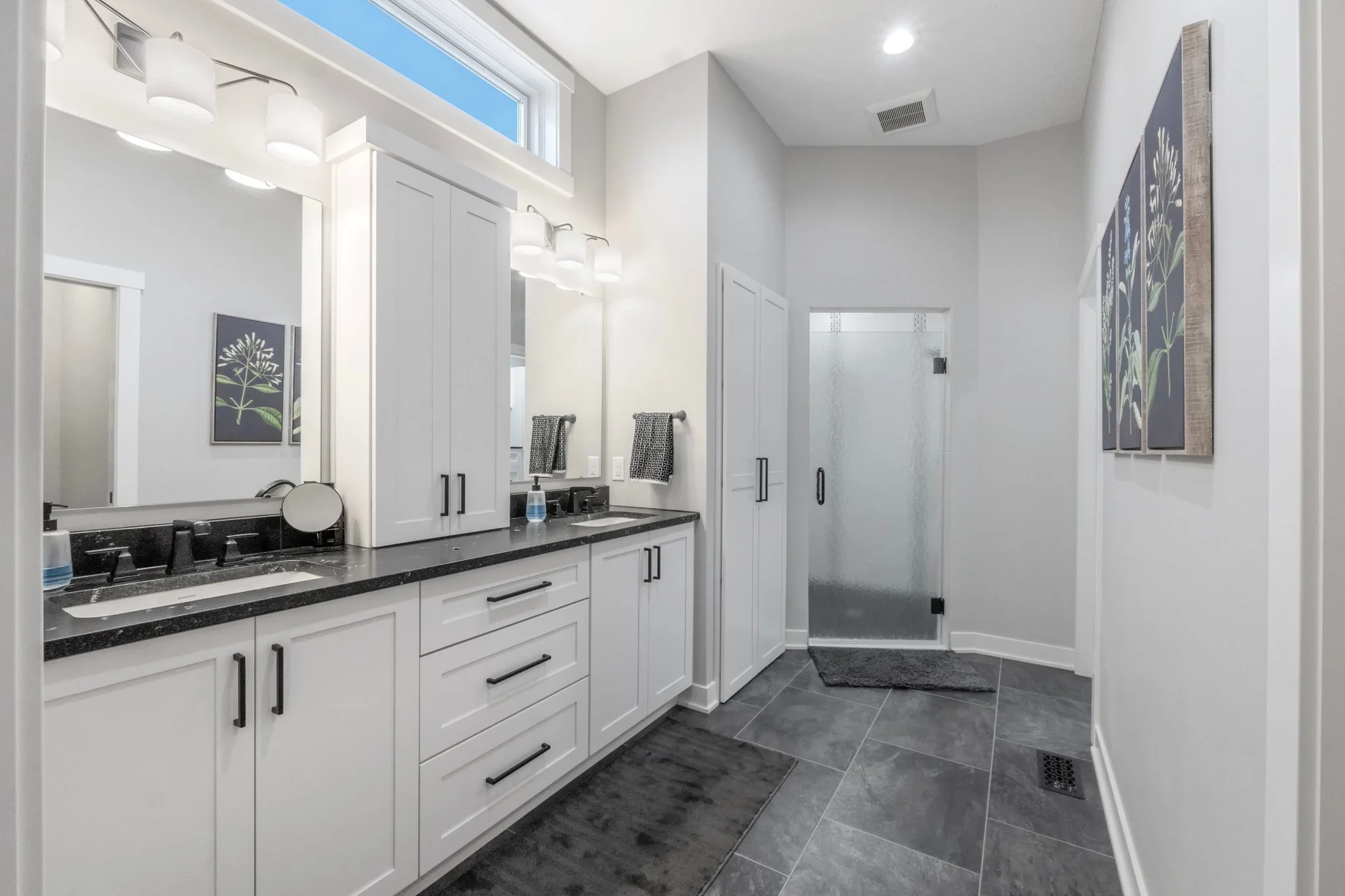 Modern bathroom with white cabinetry, black countertops, large mirrors, gray tiled floor, frosted glass shower door, and framed botanical artwork.