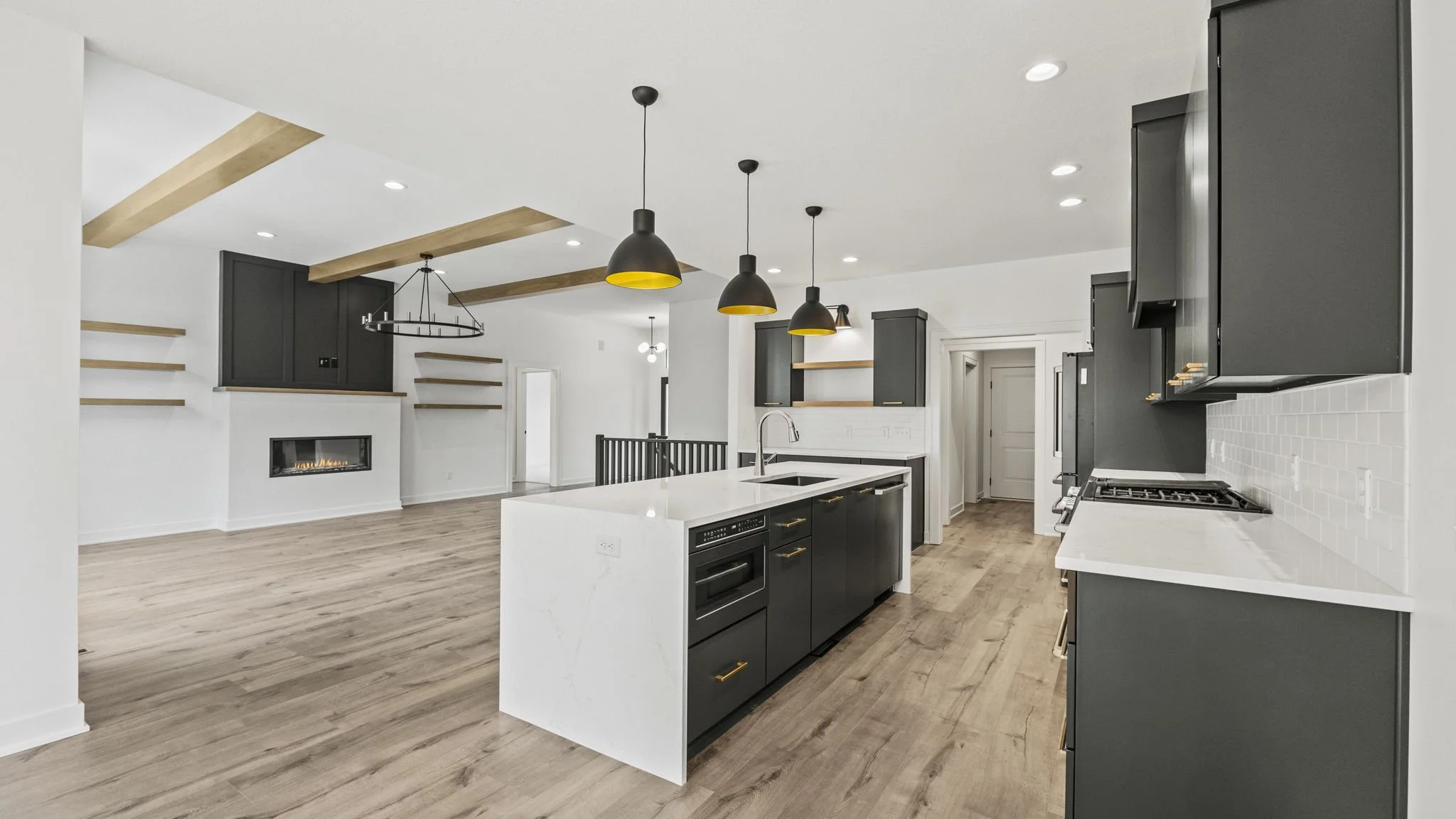 Modern open-concept kitchen with black cabinetry, white countertops, and a kitchen island with a sink. The space features black pendant lights, wood ceiling beams, and light wood flooring. The background shows a living area with a linear fireplace an