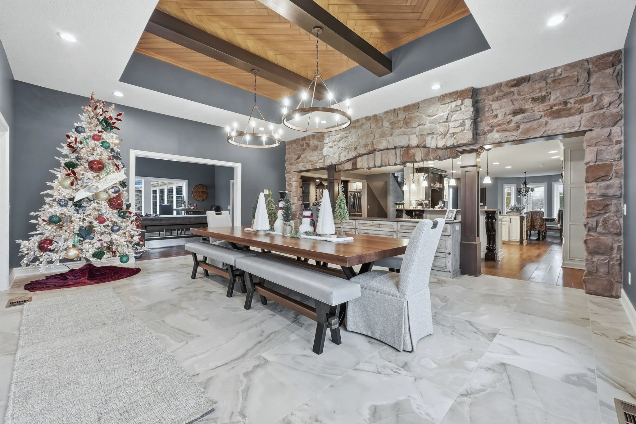 A dining room decorated for Christmas with a white tree adorned with red, gold, and blue ornaments, and a red tree skirt. The room has a large wooden dining table with a holiday centerpiece of small white trees and holiday decor. The flooring is marb