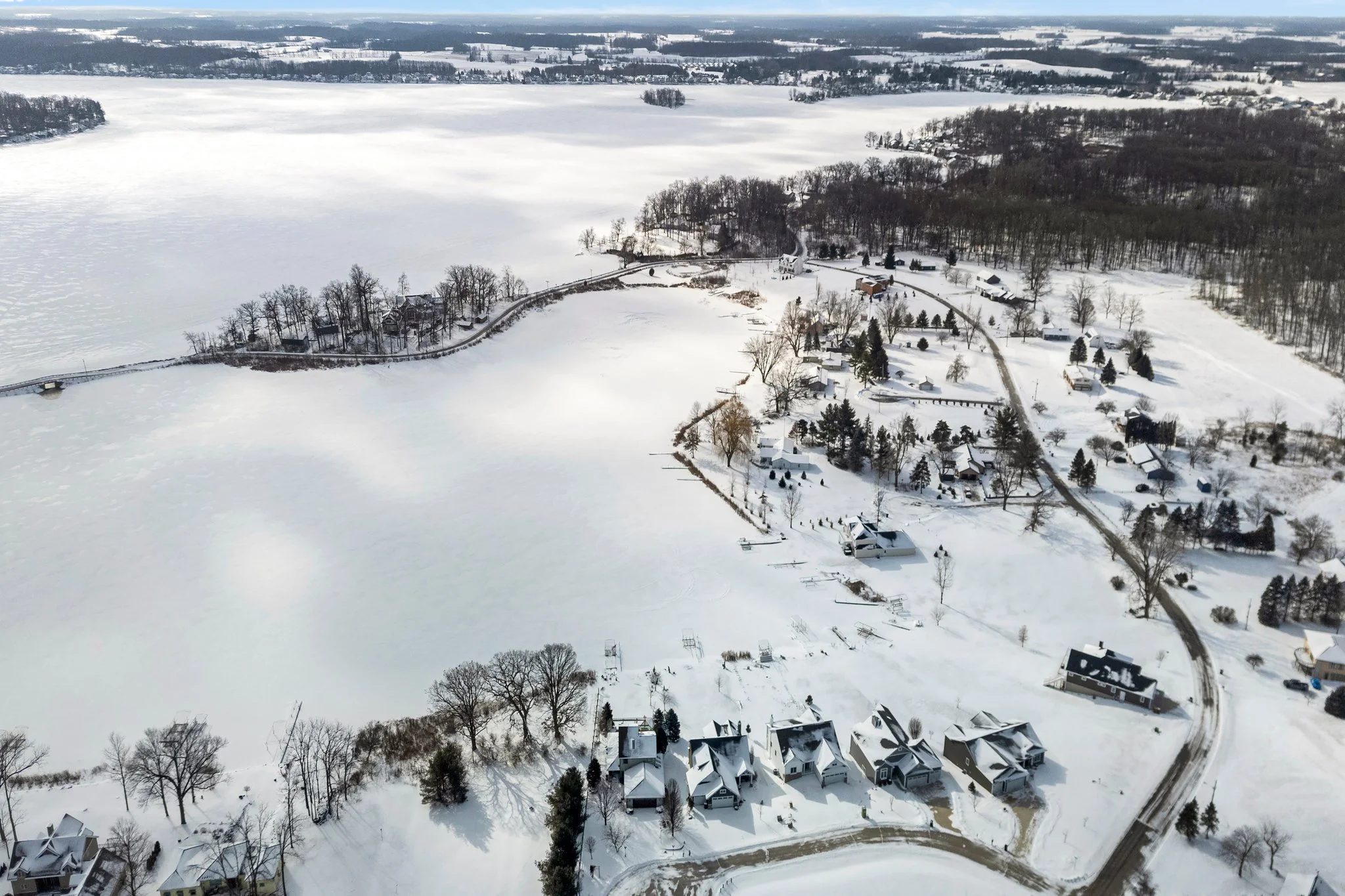 Aerial view of a snow-covered residential area with houses, trees, and roads, near a frozen lake in winter.