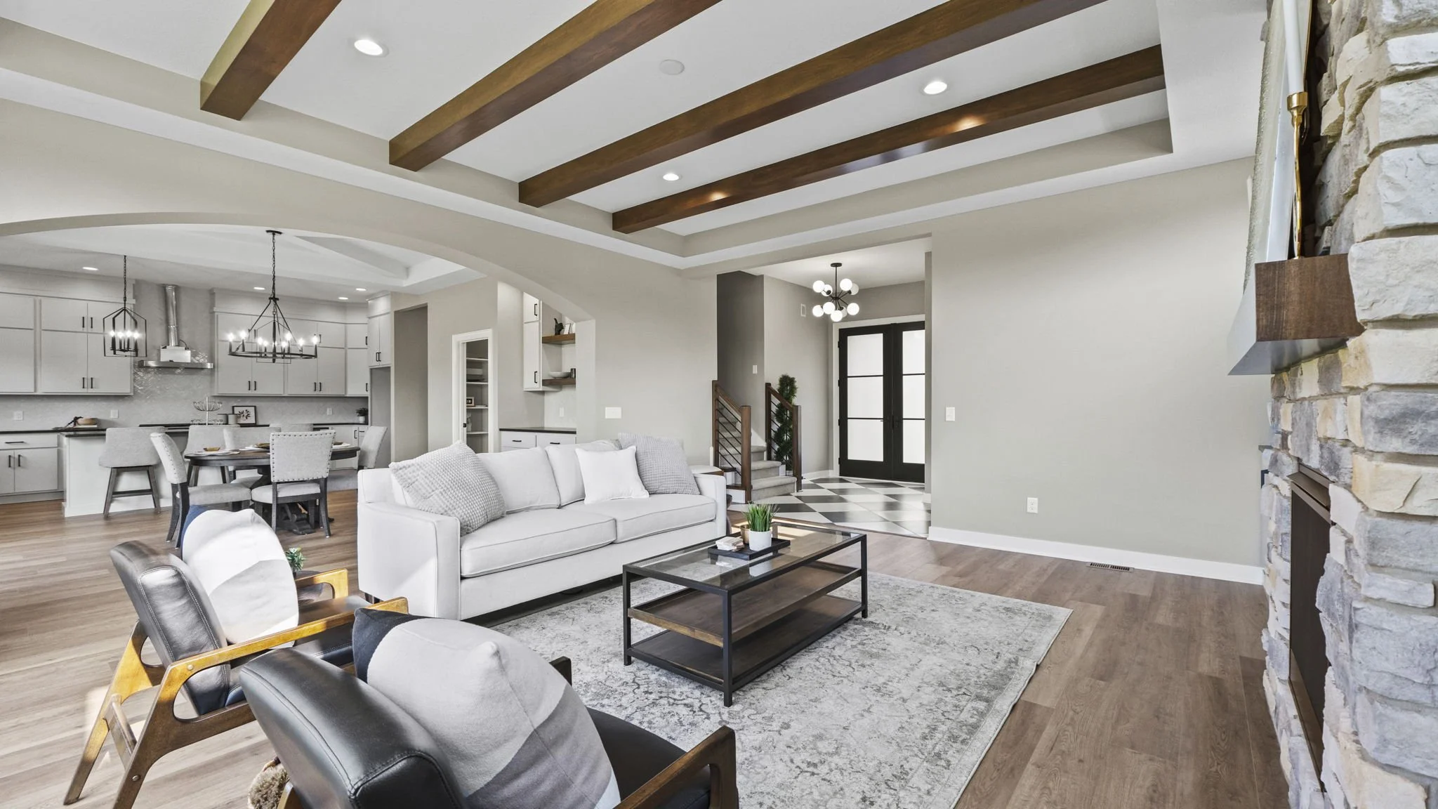 Modern living room with white sofa, black coffee table, and stone fireplace, open to a dining area and kitchen, with wooden ceiling beams, hardwood floors, and contemporary lighting.