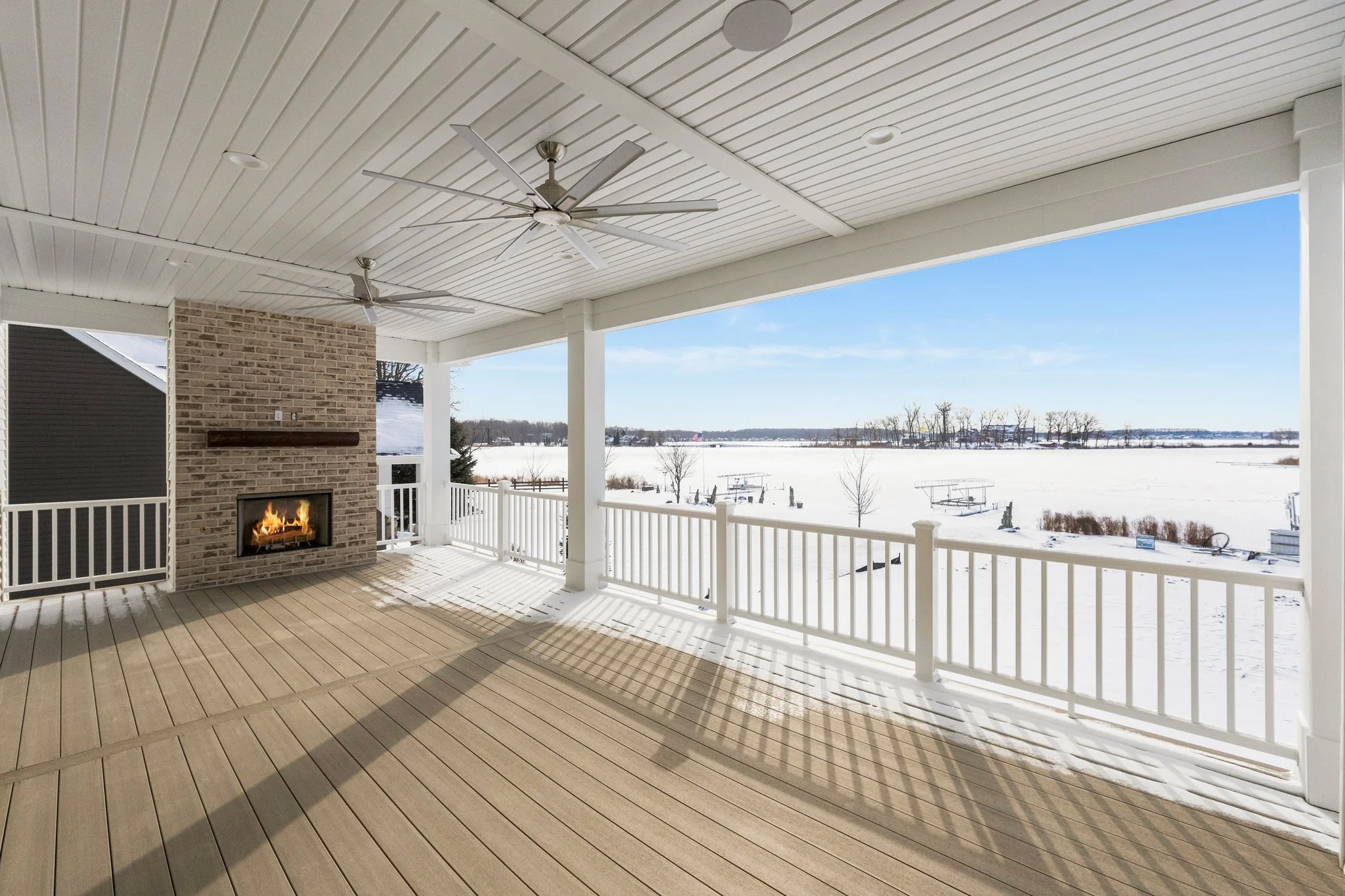 Covered porch with a fireplace, ceiling fans, and a railing overlooking a snowy lake and distant trees.