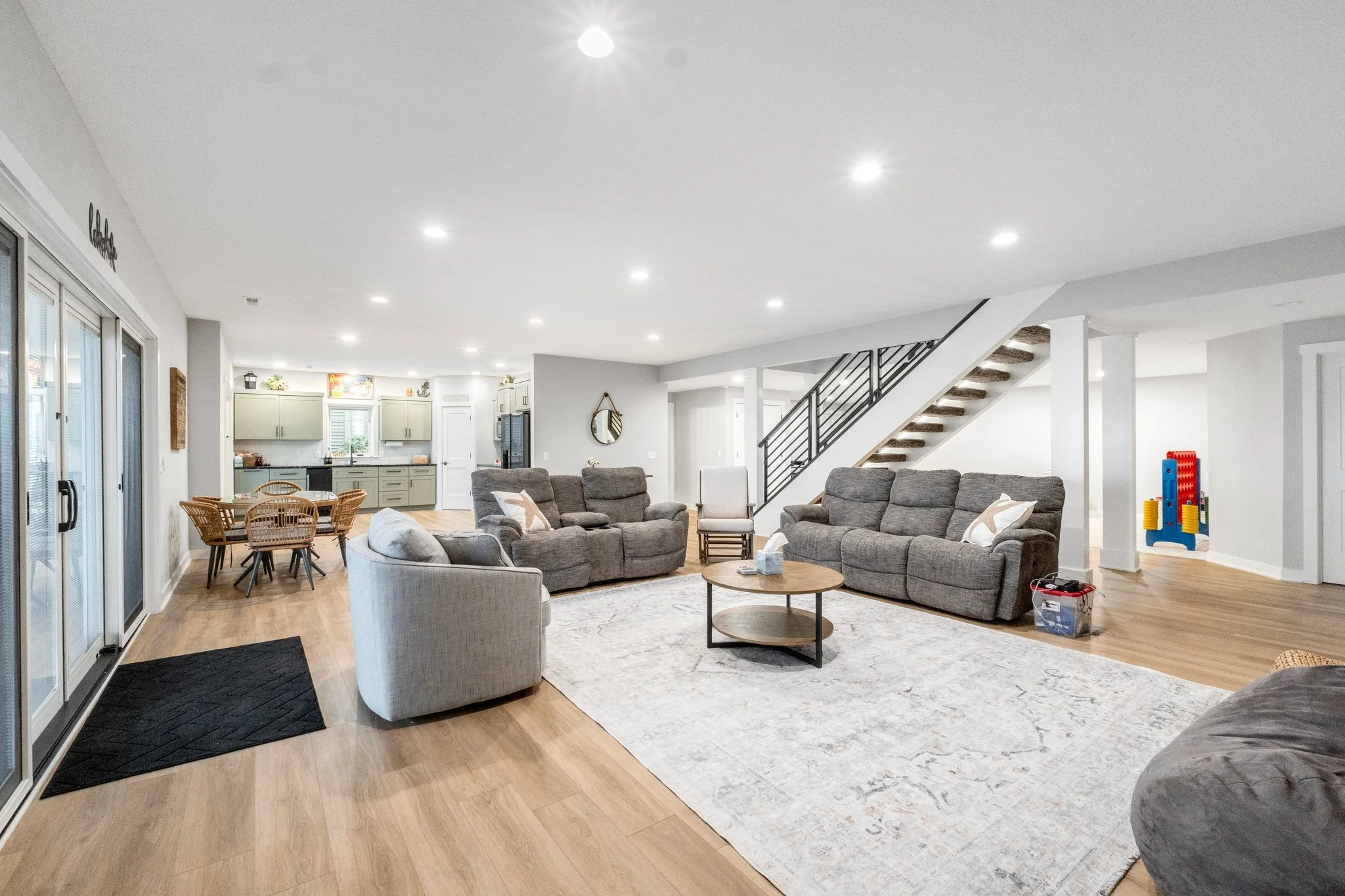 Open-concept living room with gray sofas, armchair, wooden coffee table, area rug, light wood flooring, staircase with black railing, and visible kitchen area in the background.