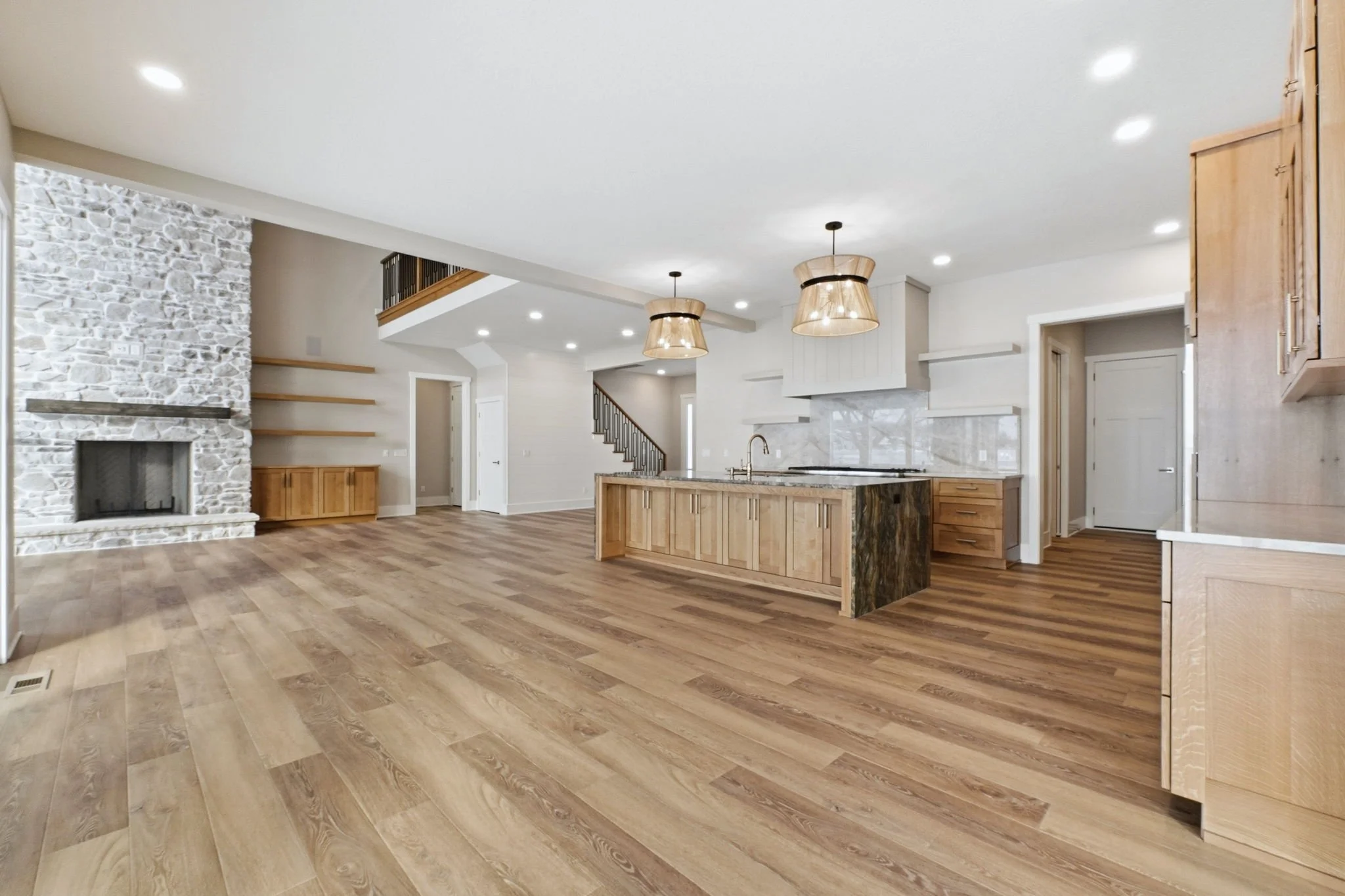 Open-concept living room and kitchen with wood flooring, a stone fireplace, wooden cabinetry, a kitchen island with a marble countertop, and modern lighting fixtures.