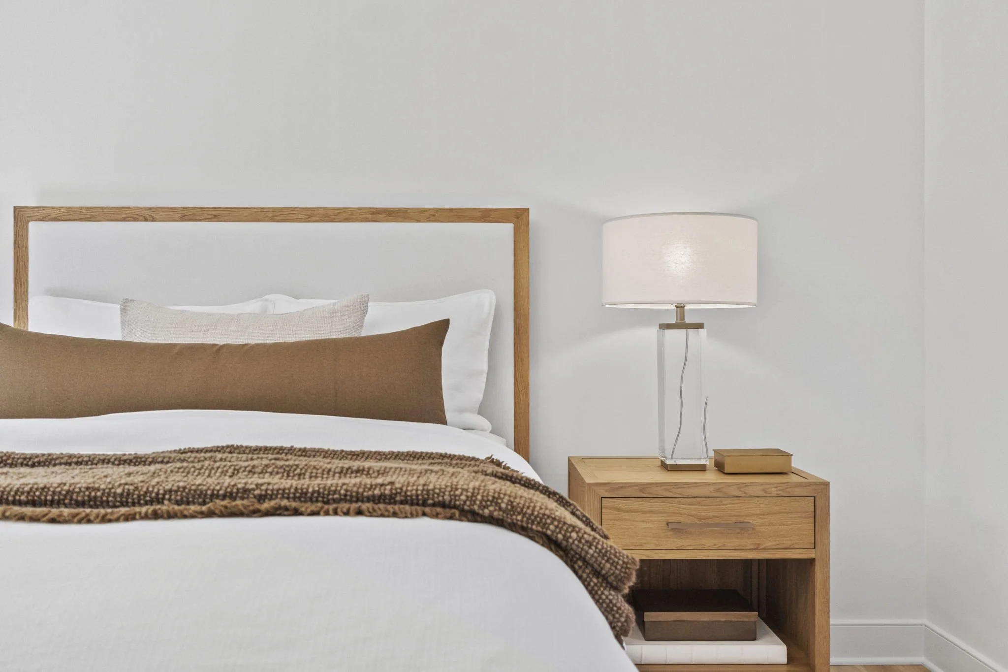 Bedroom with white walls, a wooden bed frame, white bedding, brown and beige pillows, a brown blanket, a wooden nightstand, a table lamp with a white shade, a closed book on the nightstand.