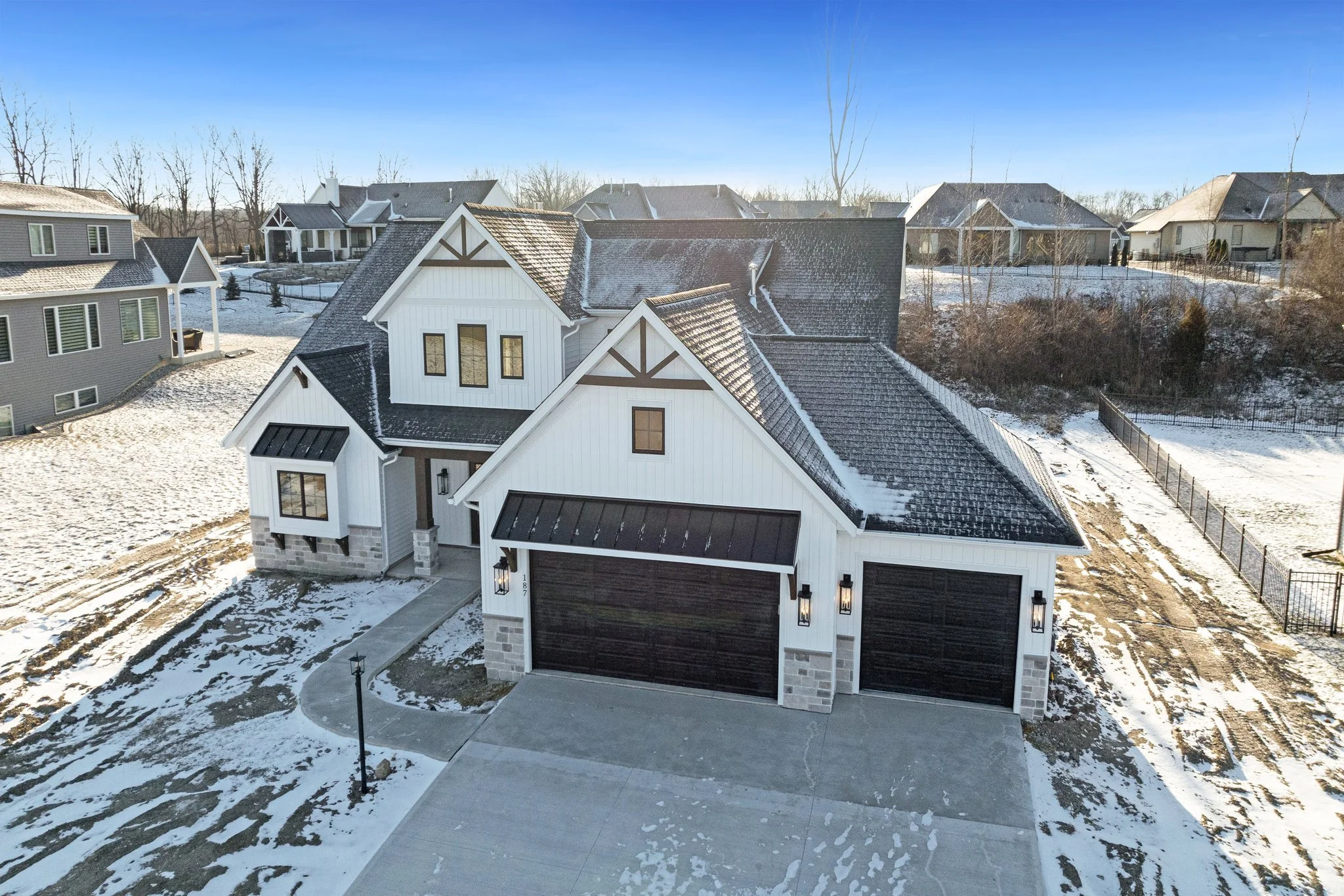 A modern two-story house with a white exterior, black garage doors, and a black metal roof, surrounded by light snow, in a neighborhood with other houses and a blue sky.