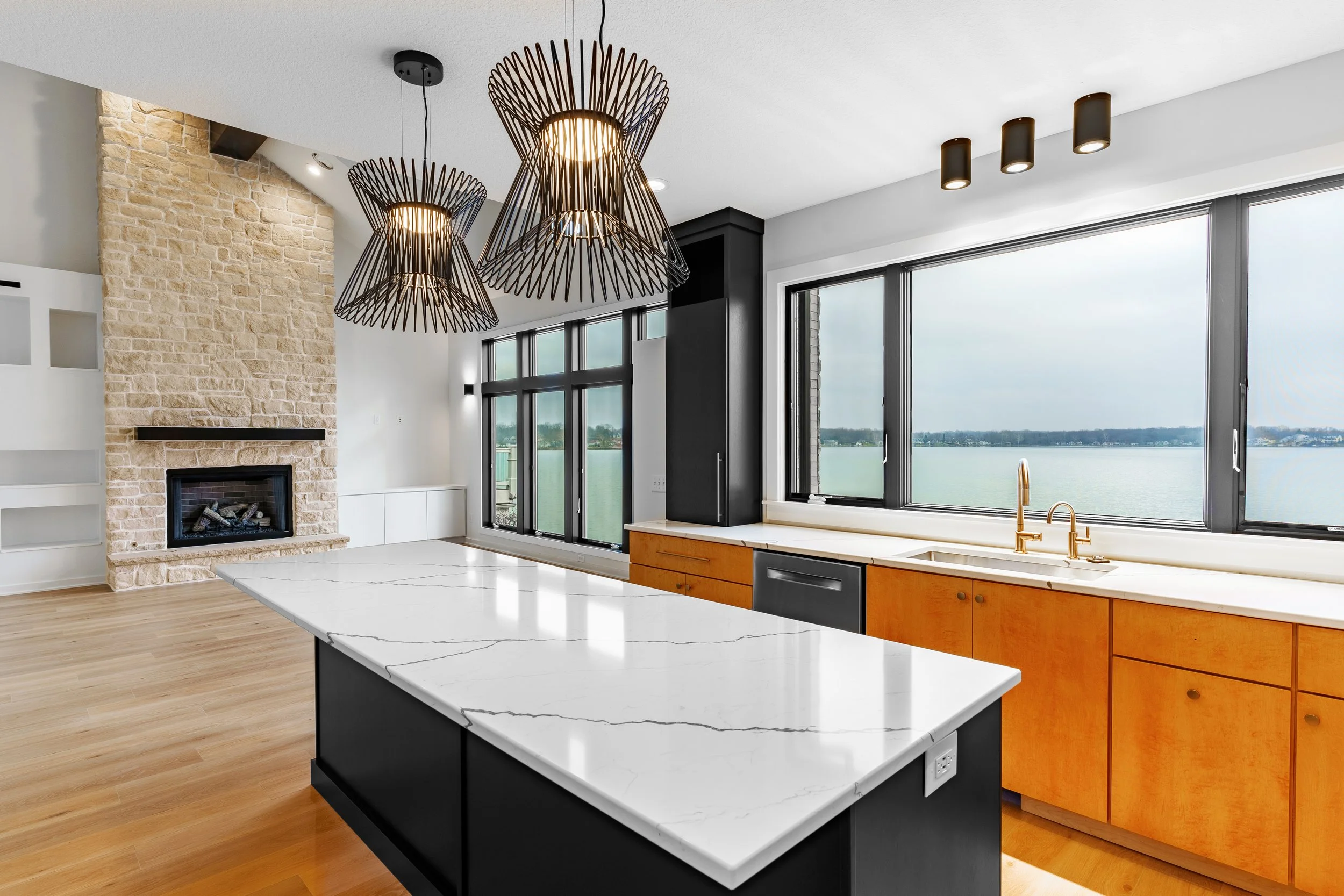 Modern kitchen with large windows overlooking a body of water, featuring a marble island, wooden cabinets, black and gold fixtures, and pendant lights with a contemporary design.