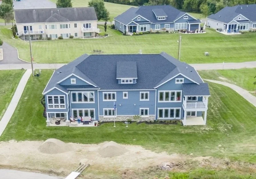 Aerial view of a large blue multi-story house with a dark roof, multiple balconies, and a well-maintained lawn, in a suburban neighborhood with other similarly styled houses.