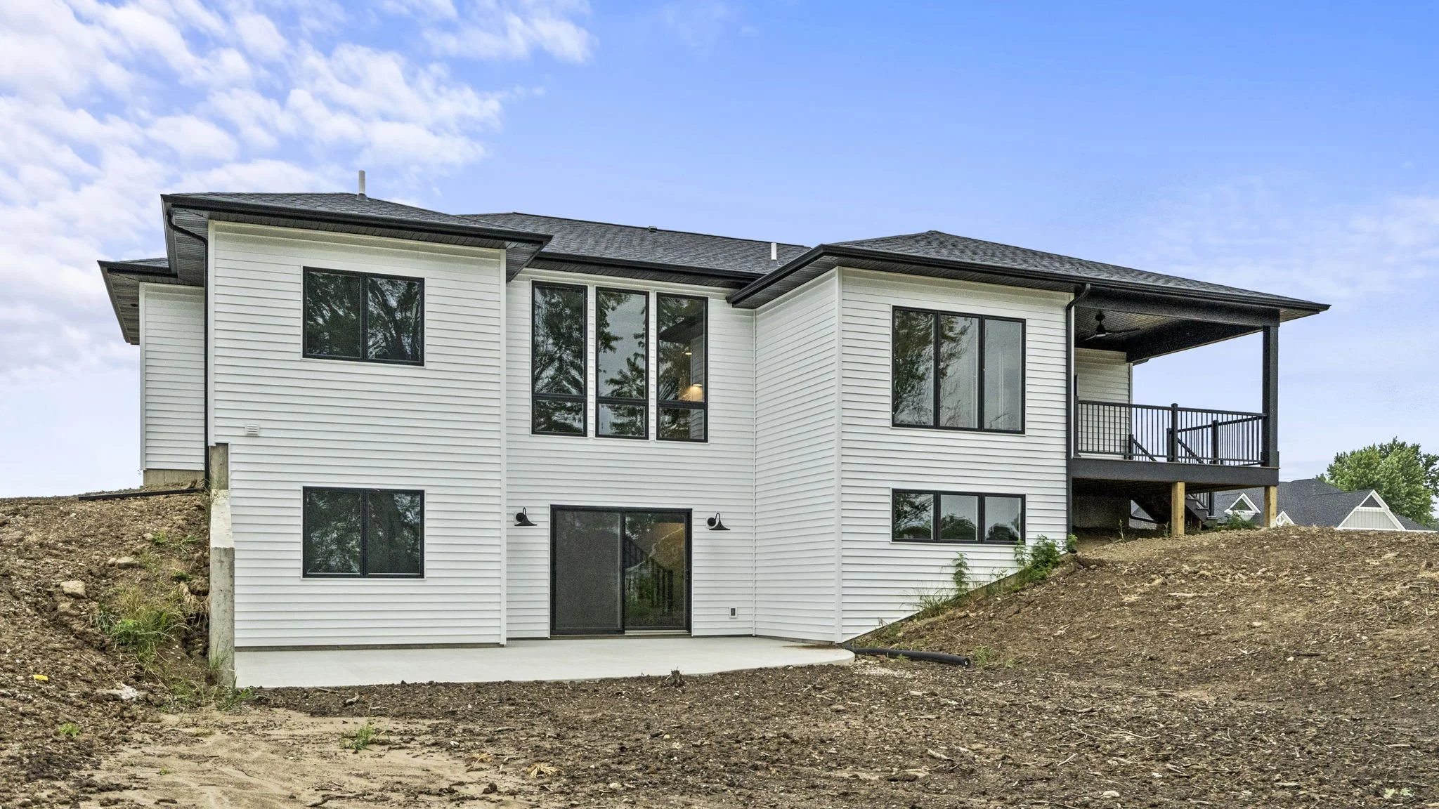 Rear view of a modern two-story house with white siding, black trims, and a small balcony, situated on a slope with exposed soil in the yard.