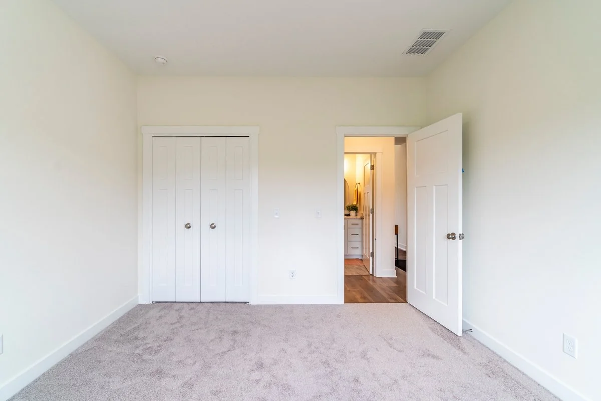 Empty bedroom with beige carpet, white walls, and a white closet door. There is an open door leading to a bathroom or hallway with hardwood floors and a vanity visible.