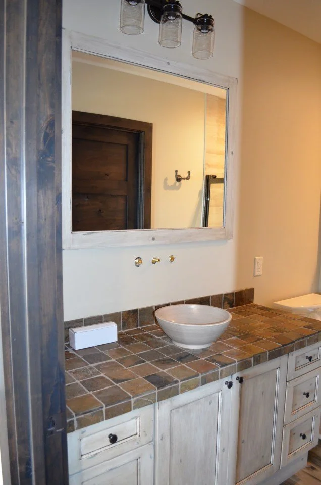 Bathroom vanity with a square mirror, a rustic faucet inset in a ceramic bowl sink, and a tiled countertop with a small box or soap holder on the left. There are three holes on the wall for additional fixtures and a ladder reflected in the mirror.
