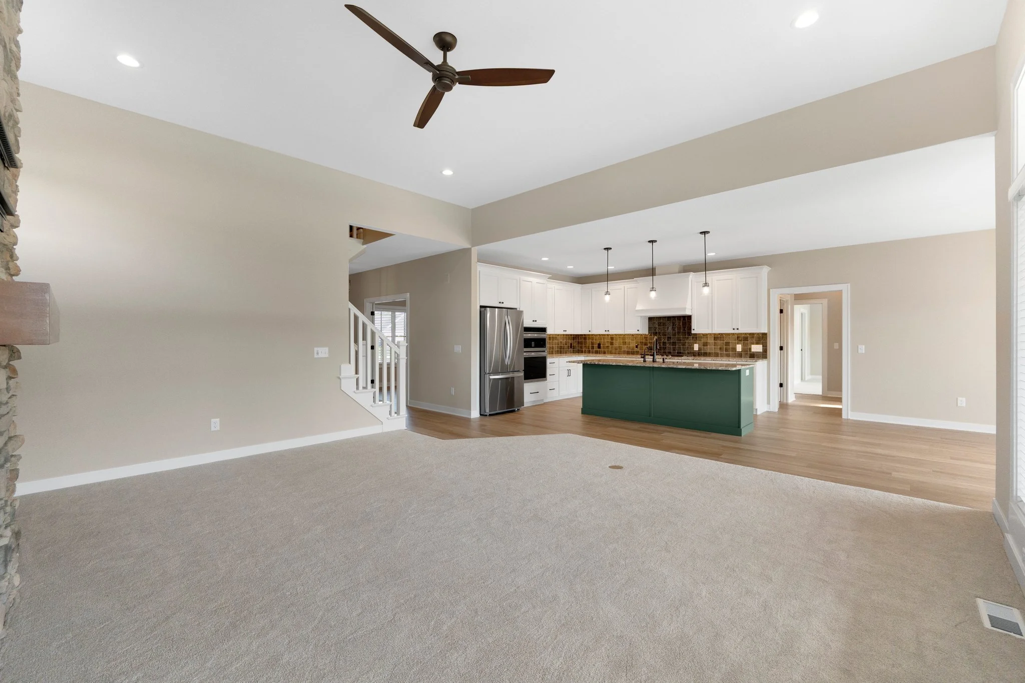 Open-concept living room and kitchen with beige walls, carpeted flooring in the living area, hardwood flooring in the kitchen, a ceiling fan, kitchen island painted green, white cabinetry, stainless steel appliances, and pendant lighting.