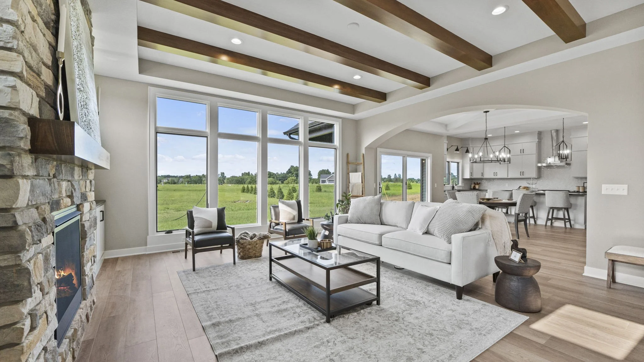 Bright living room with large windows, white sofa, two black chairs, fireplace, and view of green fields outside, adjoined to a kitchen with white cabinets and island.