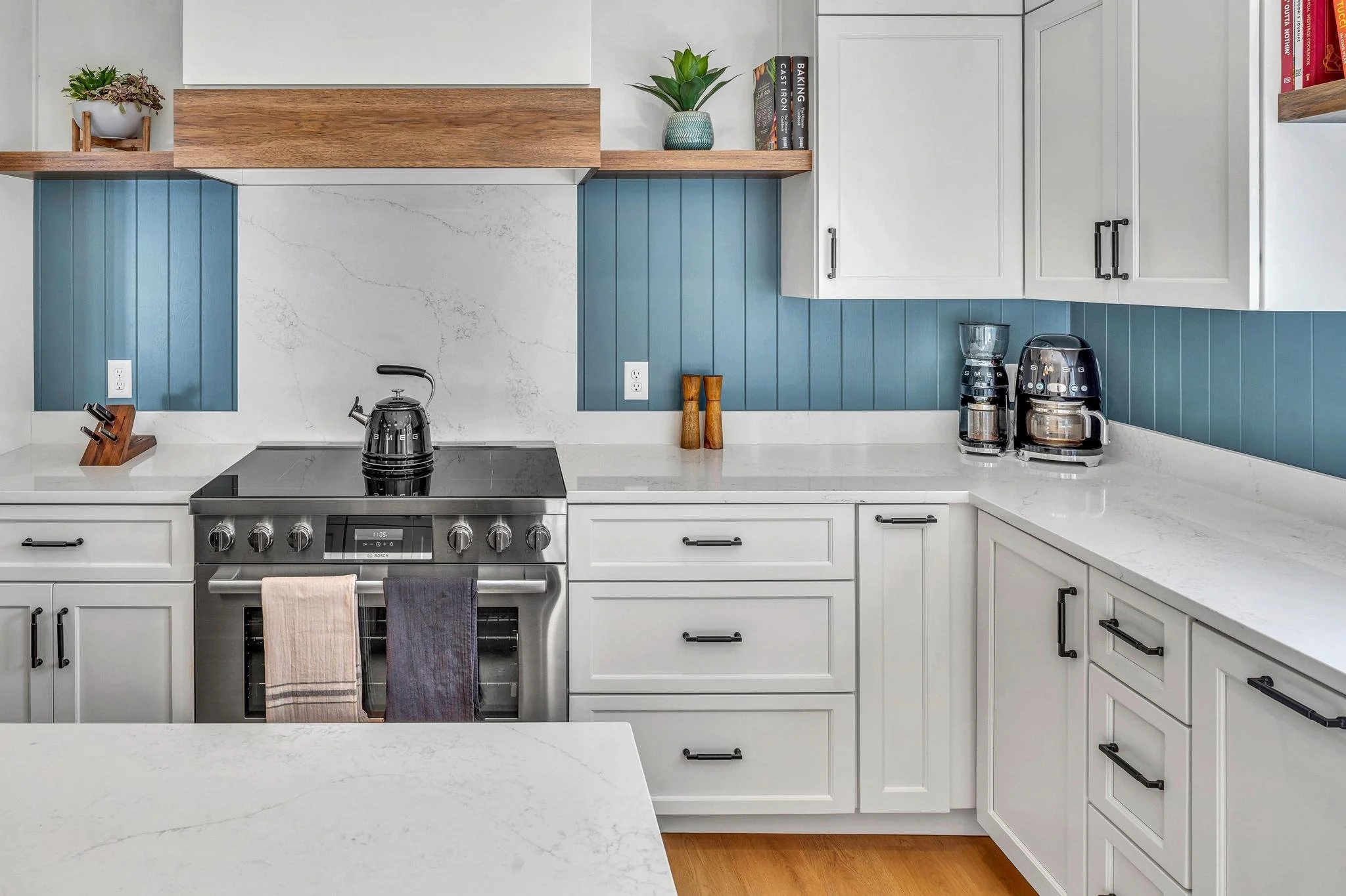Modern kitchen with white cabinets, blue wood paneling, and a stainless steel stove. Kitchen accessories include a black kettle, coffee maker, and potted plants on a wooden shelf.