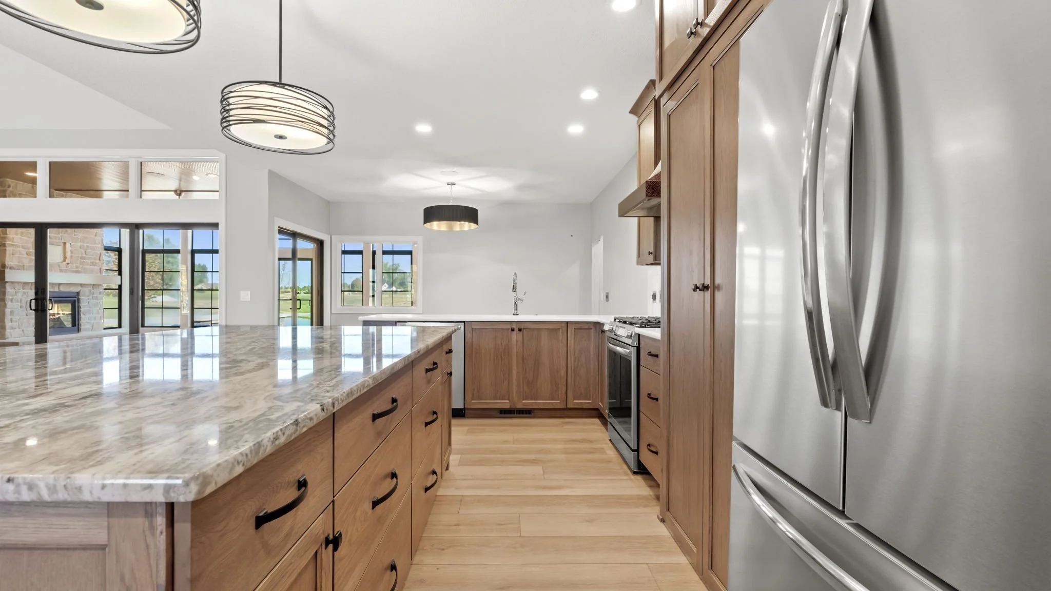 Modern kitchen with wooden cabinets, marble countertop island, stainless steel refrigerator, black oven, and bright lighting.