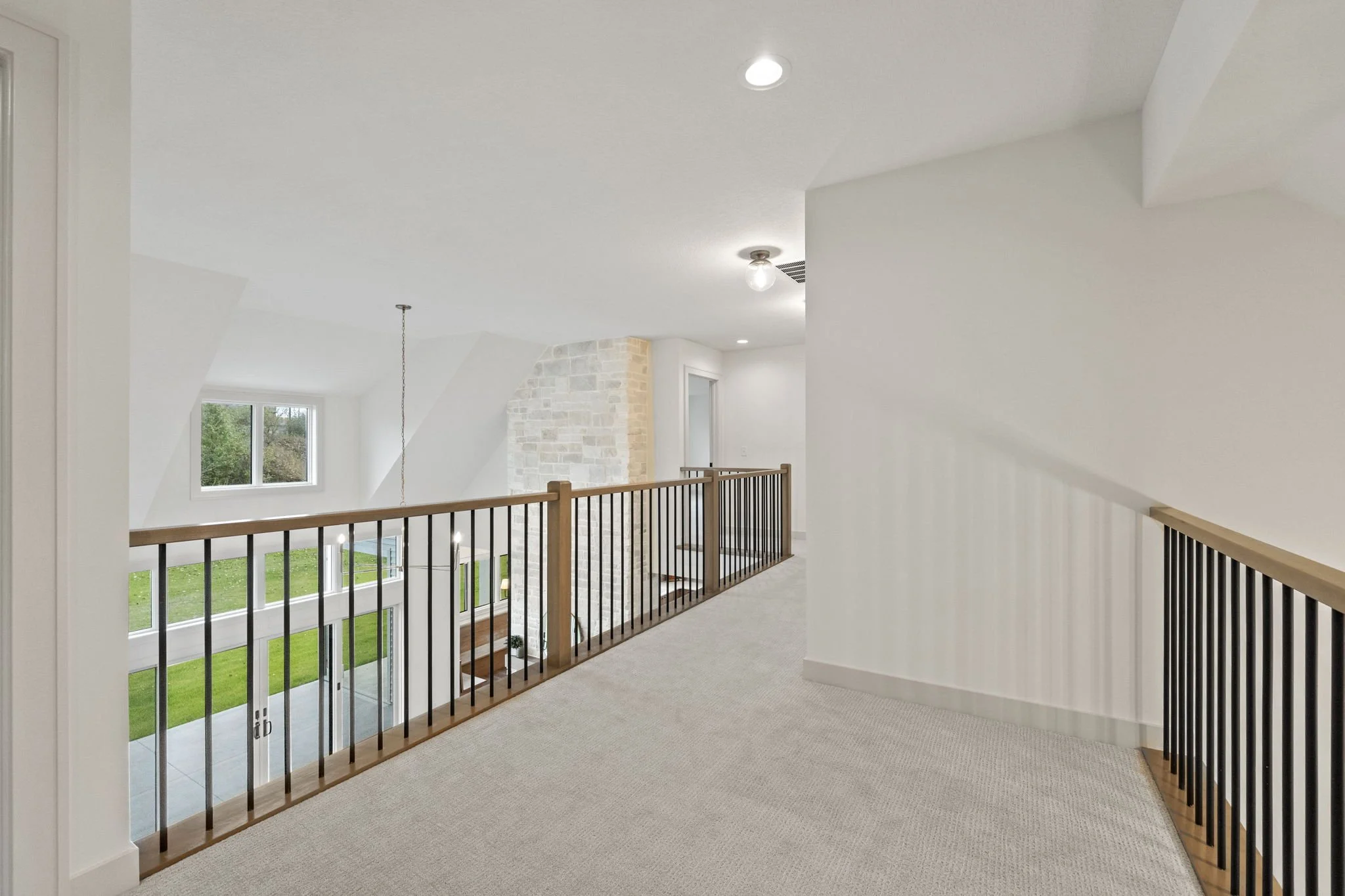 Empty indoor balcony area with white walls, beige carpet, wooden and black metal railing, ceiling lights, and a window overlooking a green yard.
