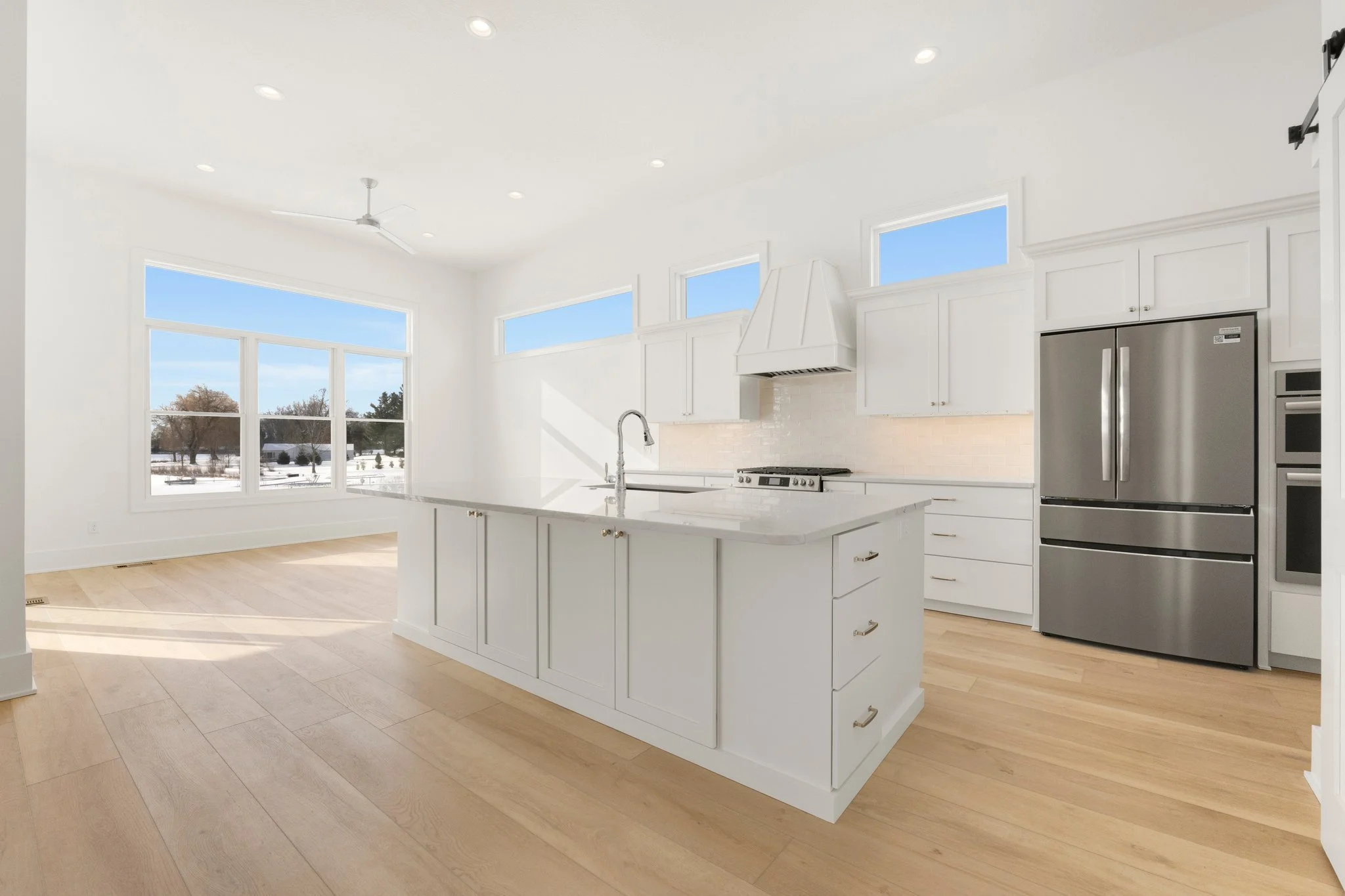 Bright, modern kitchen with white cabinets, a large island with a sink, stainless steel refrigerator, and large windows showing a winter landscape outside.