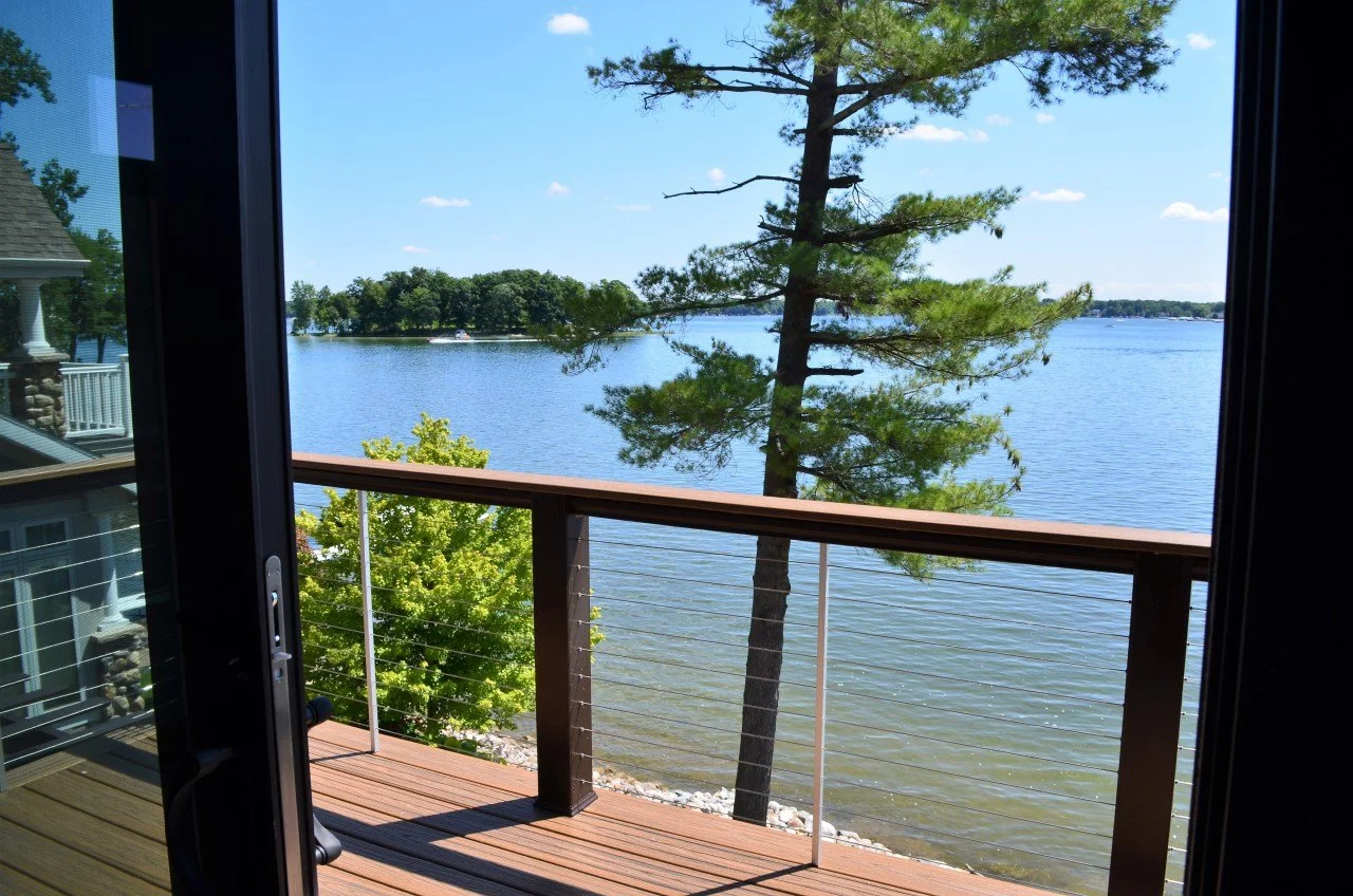 View of a lake seen from a balcony with a wooden railing, a tall pine tree, and a clear blue sky with a few small clouds.