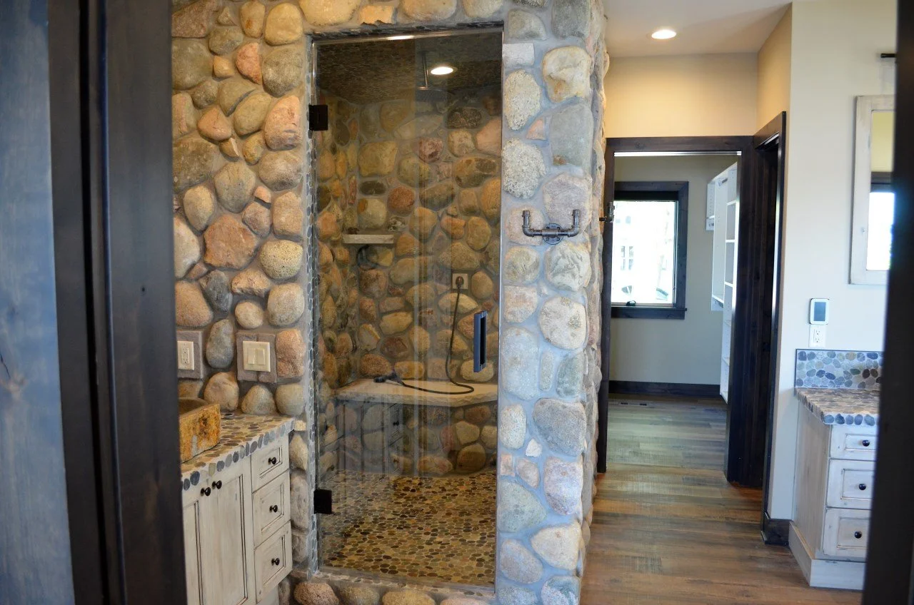 Stone shower with glass door and pebble-tile floor, adjacent to a bathroom with wooden floors and cabinets, and a window with black trim.