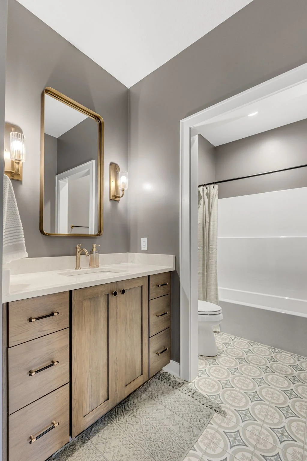Modern bathroom with gray walls, wooden vanity with brass handles, marble countertop, large mirror, wall-mounted light fixtures, and a walk-in shower with a curtain and patterned tile floor.
