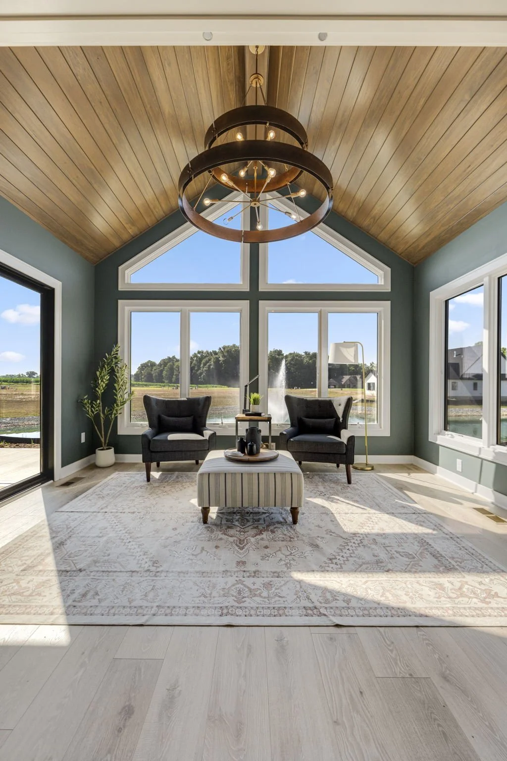 Living room with large windows, two black armchairs, a striped ottoman, a rug, potted plant, floor lamp, and a chandelier with circular design hanging from a wooden ceiling.