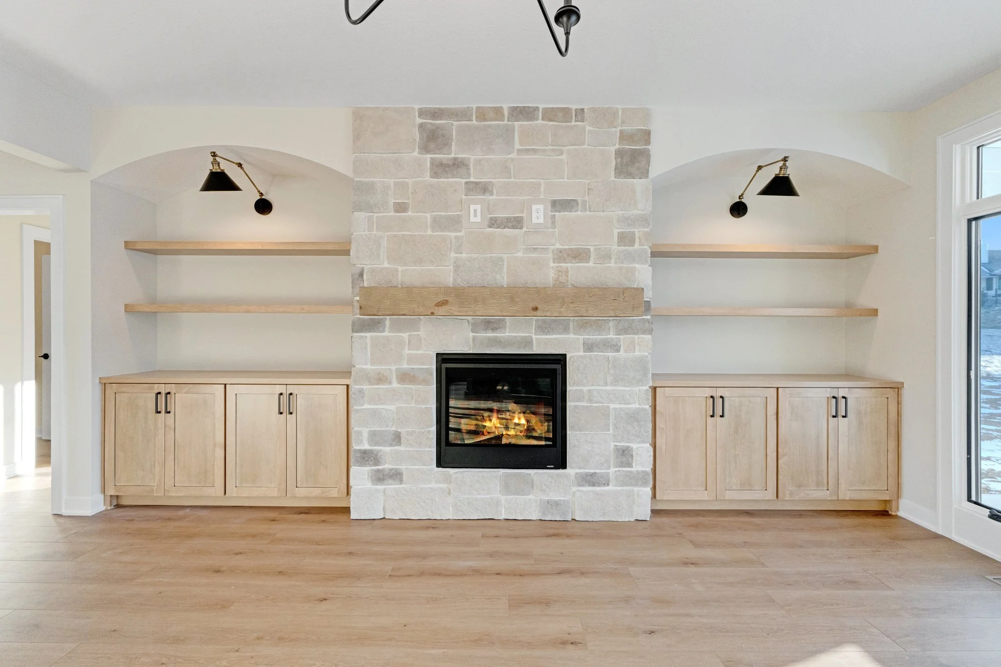 Living room with a stone fireplace, wooden built-in cabinets on both sides, open shelves above, and black wall-mounted lamps, with hardwood flooring and a large window on the right.