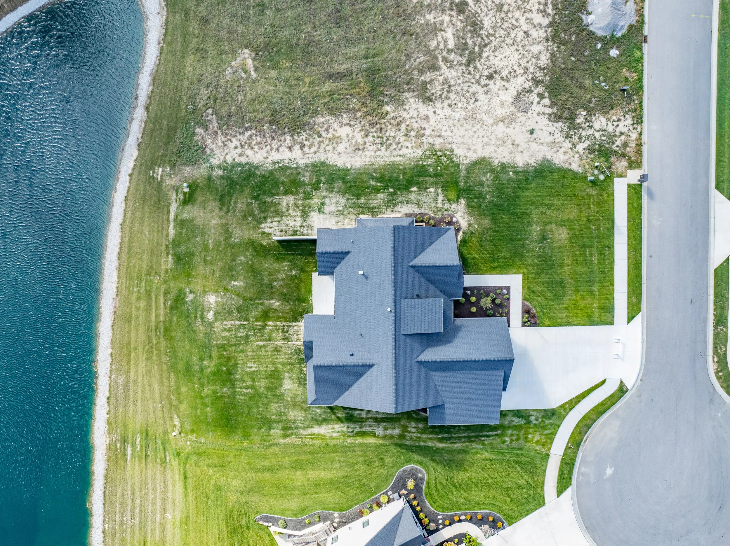 Top-down aerial view of a house with a gray roof, surrounding a well-maintained lawn, concrete driveway, and a large body of water on the left side.