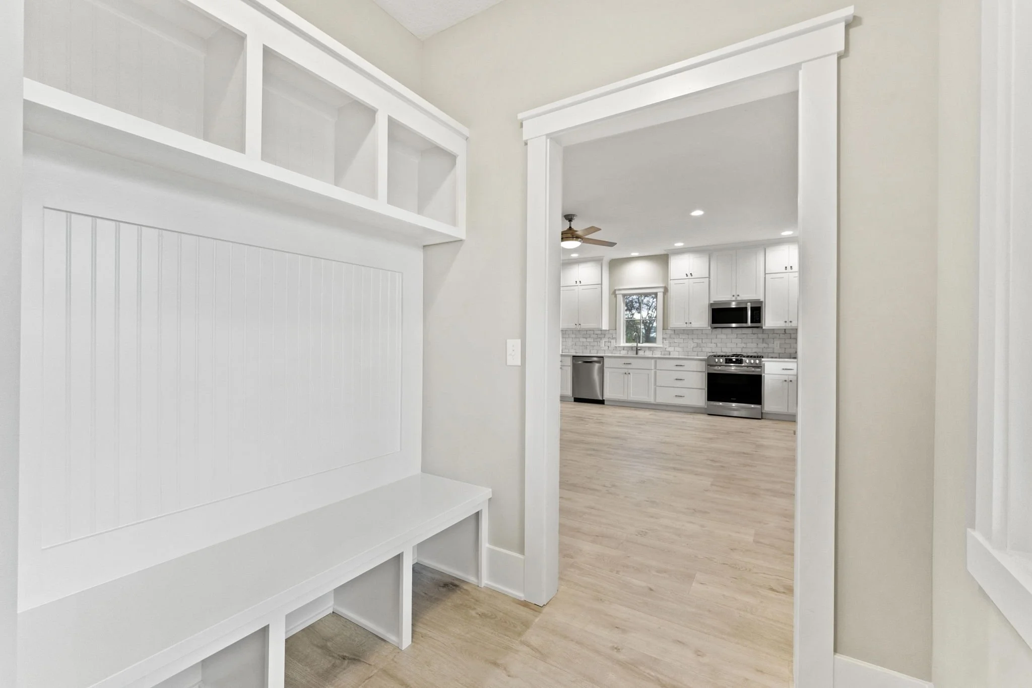 View from a hallway into a kitchen with white cabinets, a stainless steel dishwasher, stove, microwave, and a small window, with light-colored hardwood flooring.
