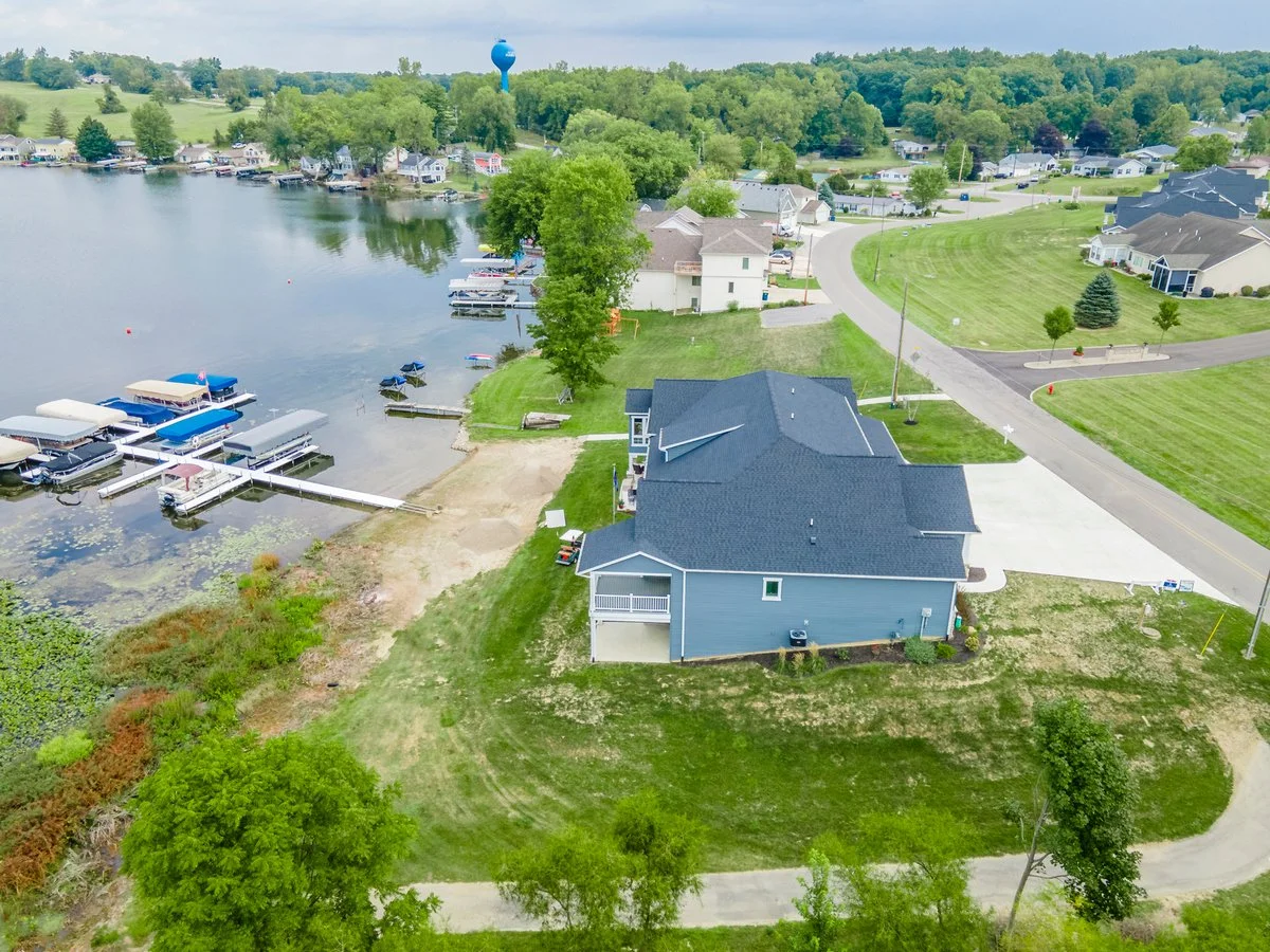 Aerial view of a lakeside residential area with houses, docks, boats, and green yards, surrounded by trees and a water tower in the background.