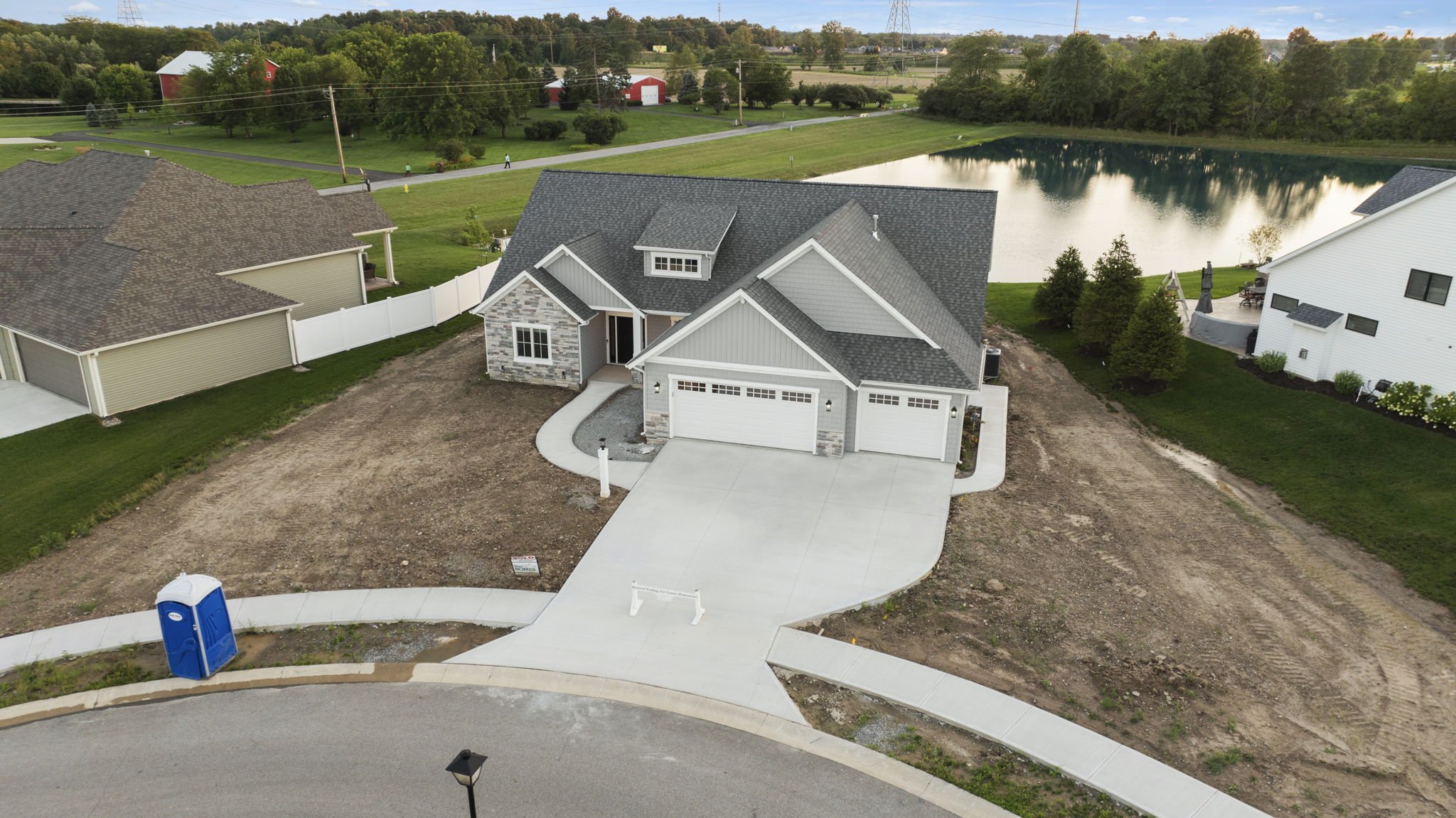 A newly built house with a gray roof and stone accents, a concrete driveway, and an empty front yard, situated in a suburban neighborhood near a pond, with neighboring houses and green trees in the background.