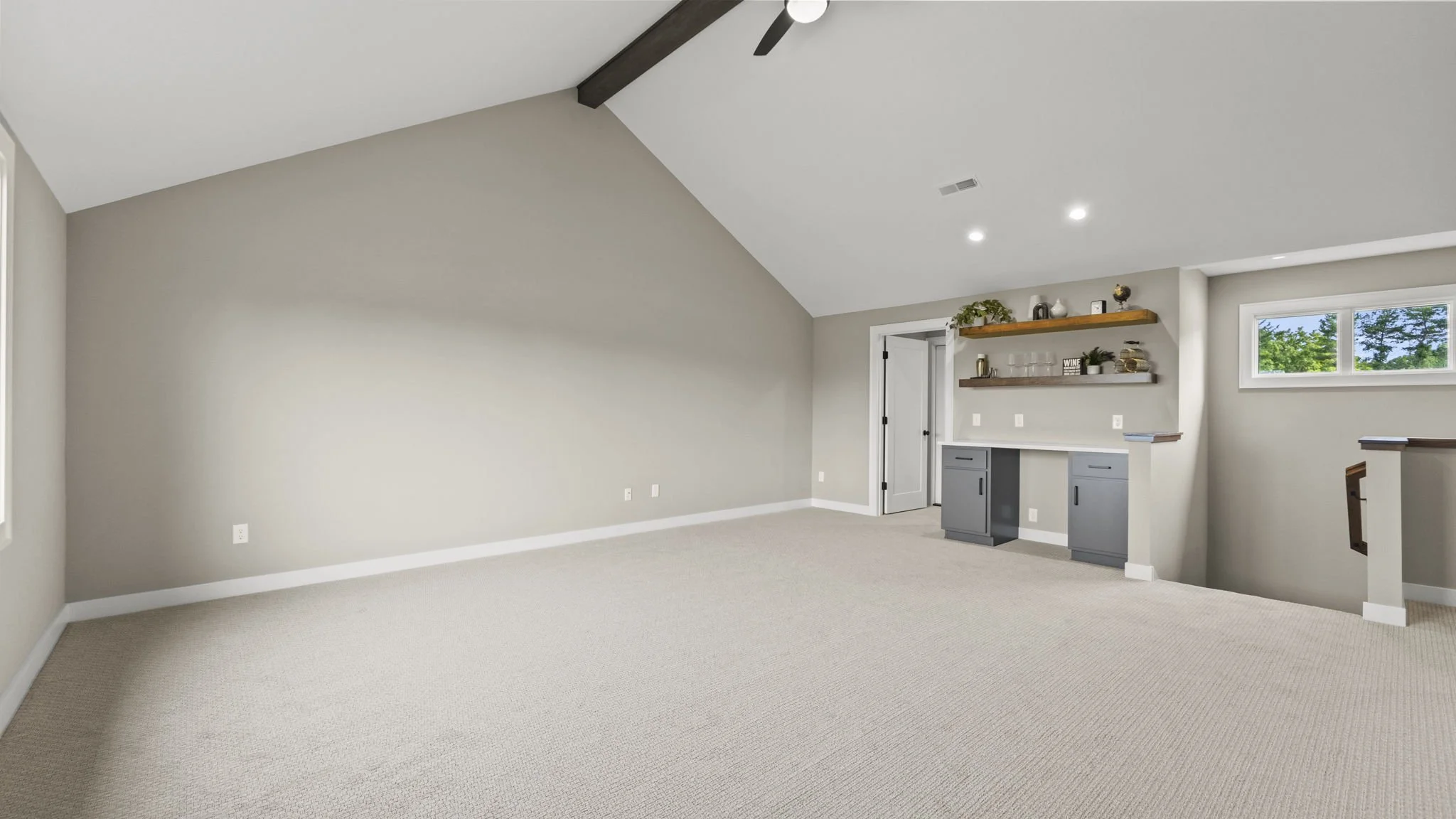 Empty living room with beige carpet, beige walls, a vaulted ceiling with a dark wooden beam, a ceiling fan, recessed lighting, a small horizontal window, and a built-in bar area with gray cabinets and wooden shelves decorated with plants and glasswar