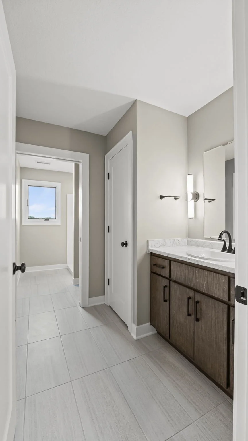 A modern bathroom with a wooden vanity, marble countertop, white sink, black faucet, large mirror, wall-mounted light, small window, and light-colored tiled floor.