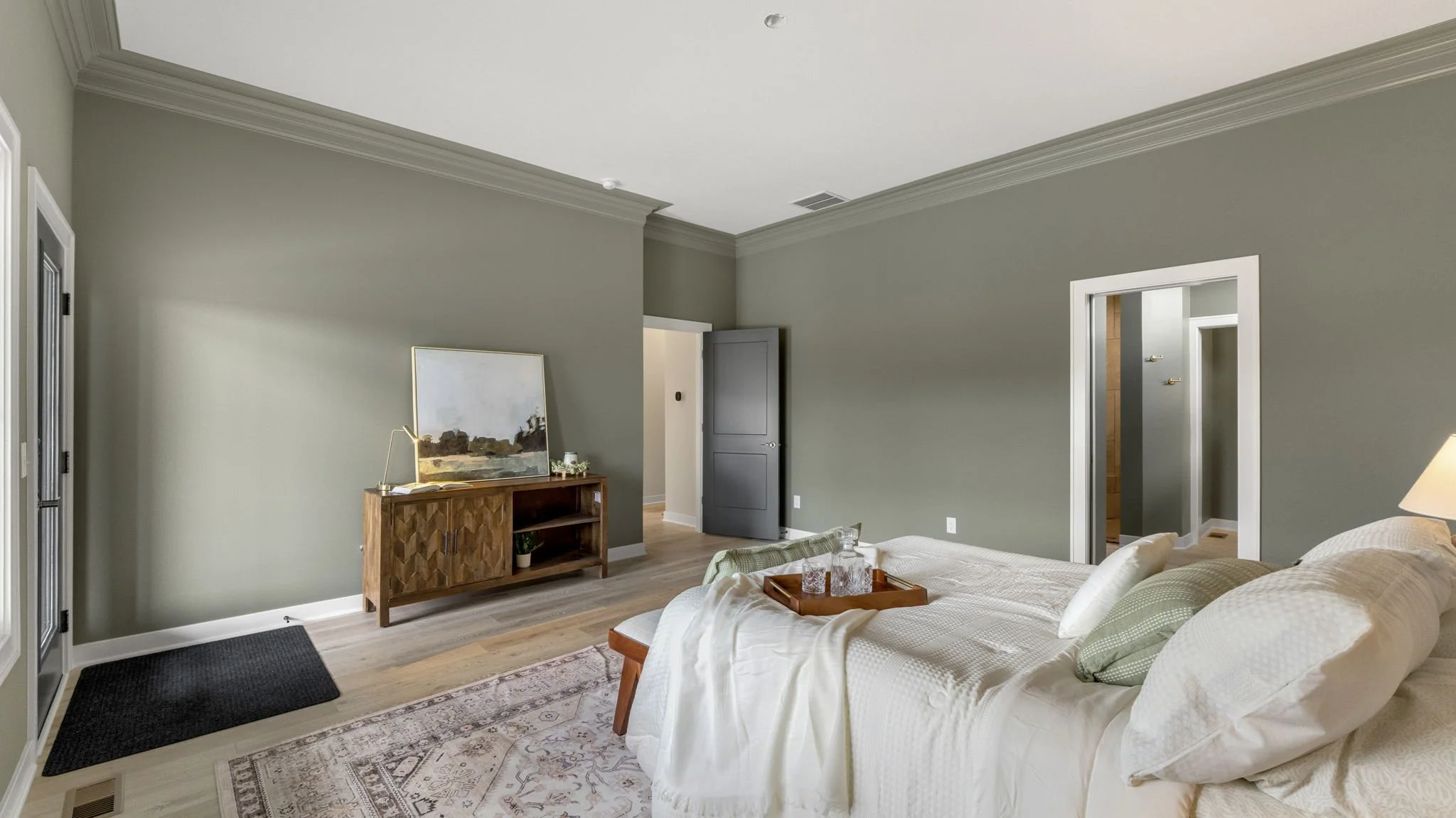 A bedroom with a bed, pillows, and blankets, a wooden dresser with a mirror, a black door, an open door leading to a hallway, a lamp, and a patterned rug on wooden floors.