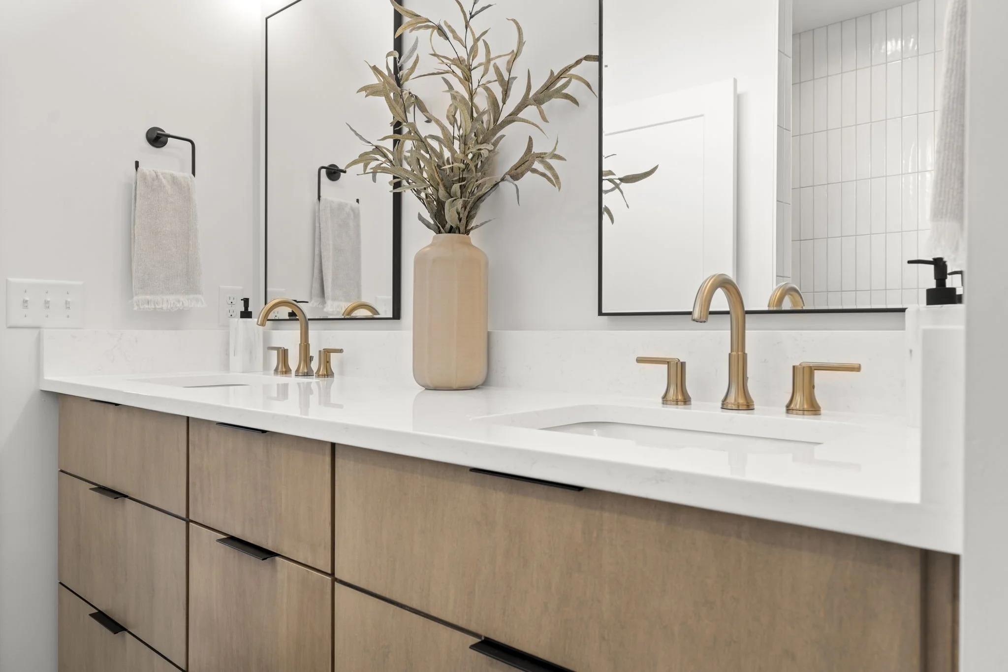 Modern bathroom vanity with a white countertop, two gold fixtures, a large beige vase with dried leaves, large mirrors, and white towels hanging on black hooks.