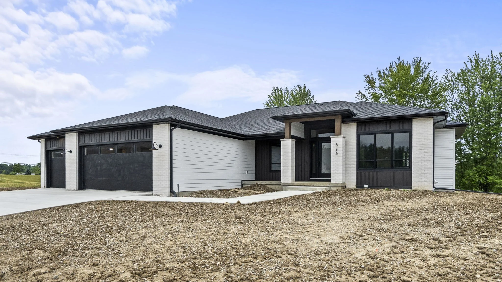 Newly constructed modern single-story house with a black and white exterior, attached two-car garage, and a small front porch, situated on a slightly sloped lot with dirt and gravel ground, surrounded by green trees under a partly cloudy sky.