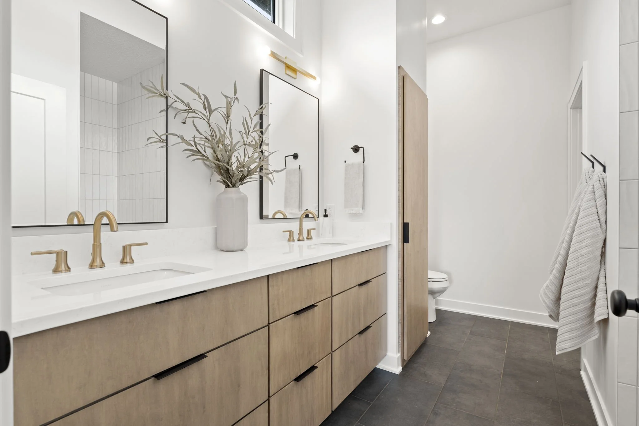 Modern bathroom with double vanity featuring wood cabinets, white countertop, gold fixtures, two mirrors, a vase with greenery, white walls, towel hooks with towels, and a toilet in the background.