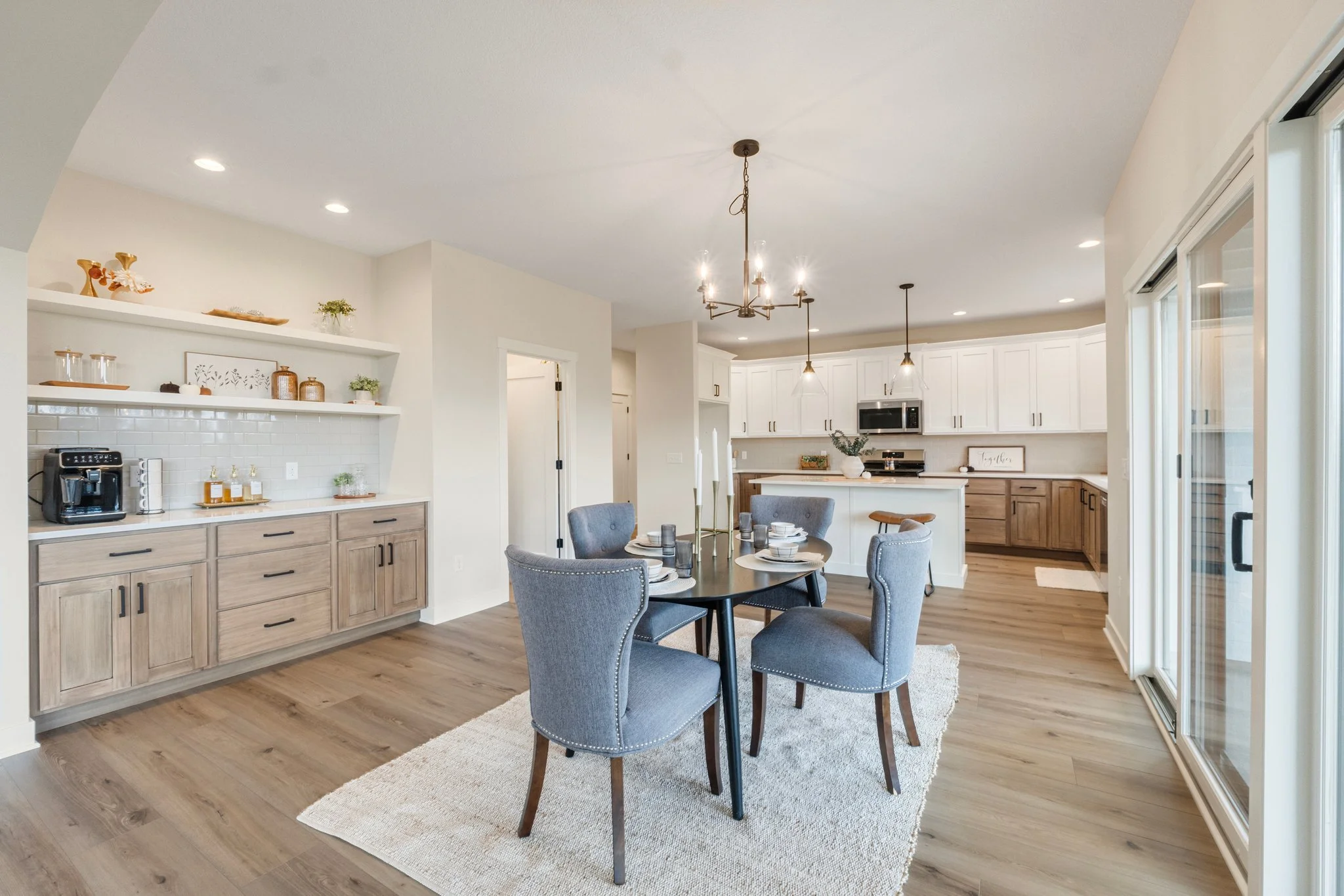 Open-concept kitchen and dining area with a round table set for four, gray upholstered chairs, white cabinetry, wood accents, and sliding glass doors to the outside.