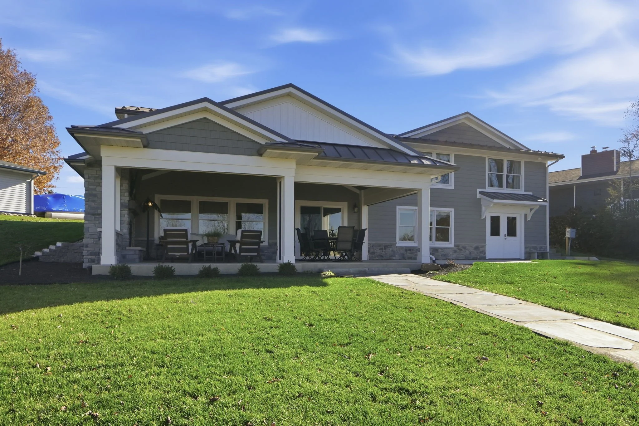 Modern two-story house with a gray exterior, white trim, and a metal roof, featuring a covered porch with outdoor furniture, a lush green lawn, and a stone pathway leading to the front entrance.