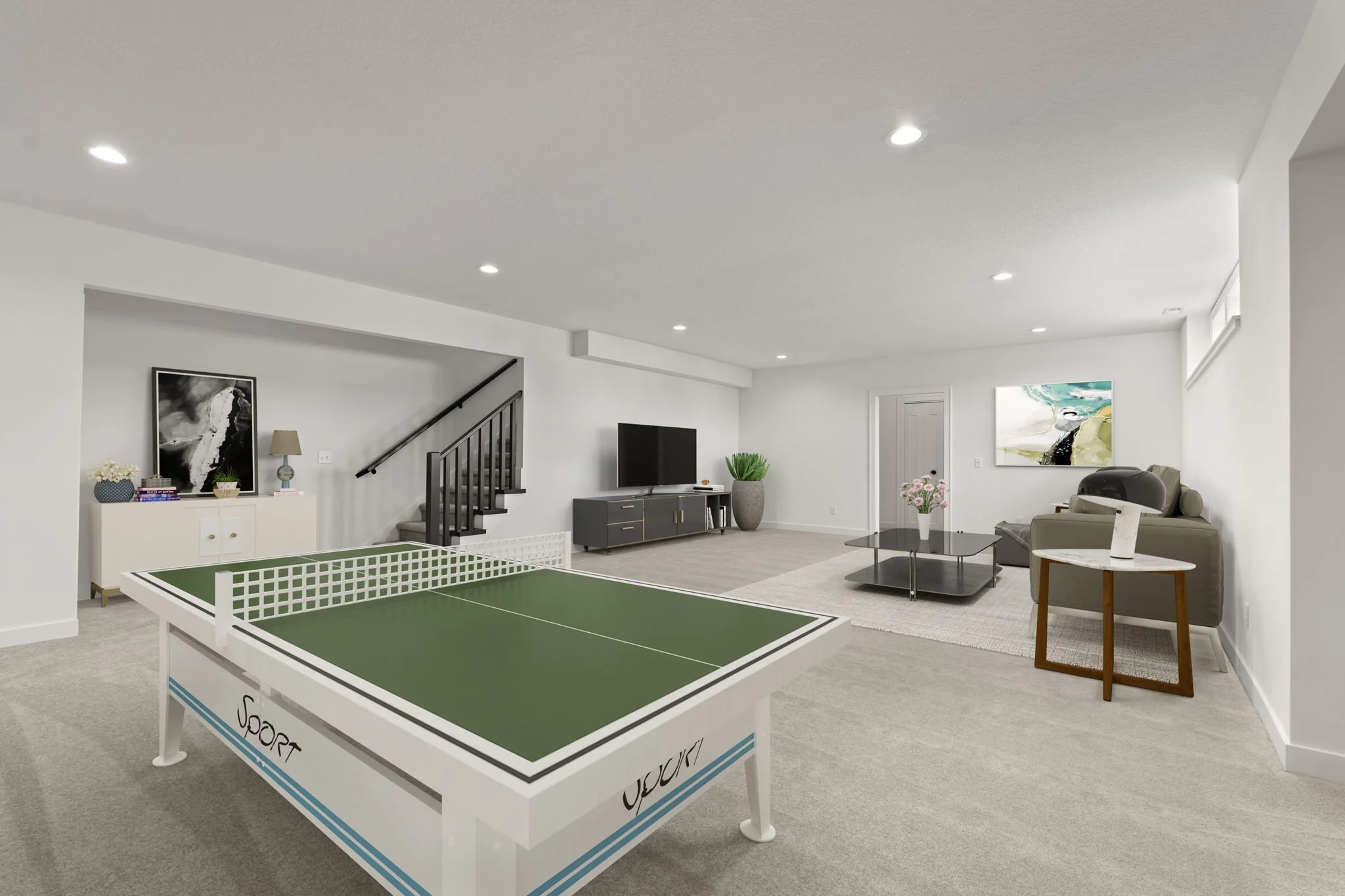 Spacious basement with a ping pong table, a TV on a stand, a gray sofa, a coffee table with flowers, a side table with a lamp, and walls decorated with artwork.