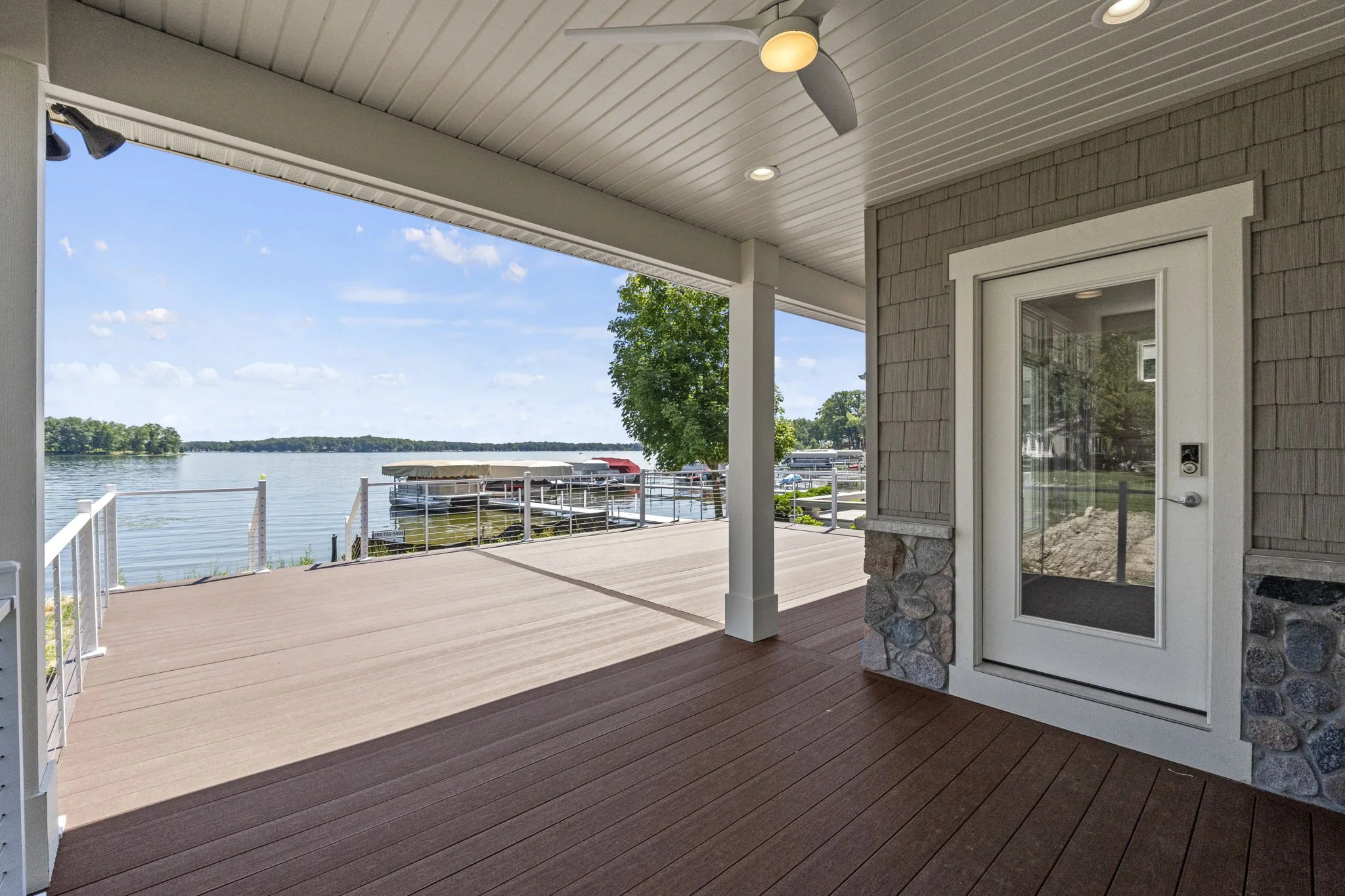 View of a lakeside porch with wooden flooring, railing, and a door, overlooking a lake with boats and a dock, under a partly cloudy sky.