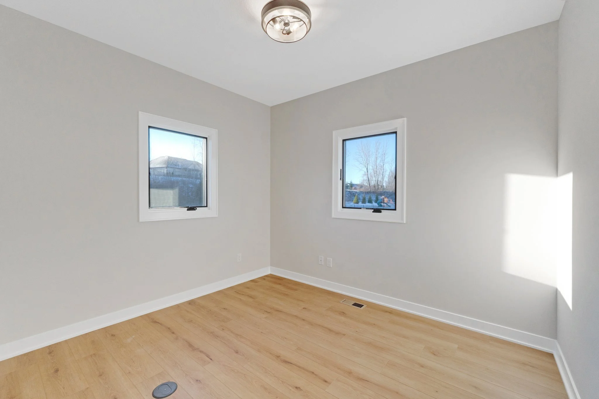 Empty room with light-colored walls, two windows, hardwood floor, and ceiling light fixture.
