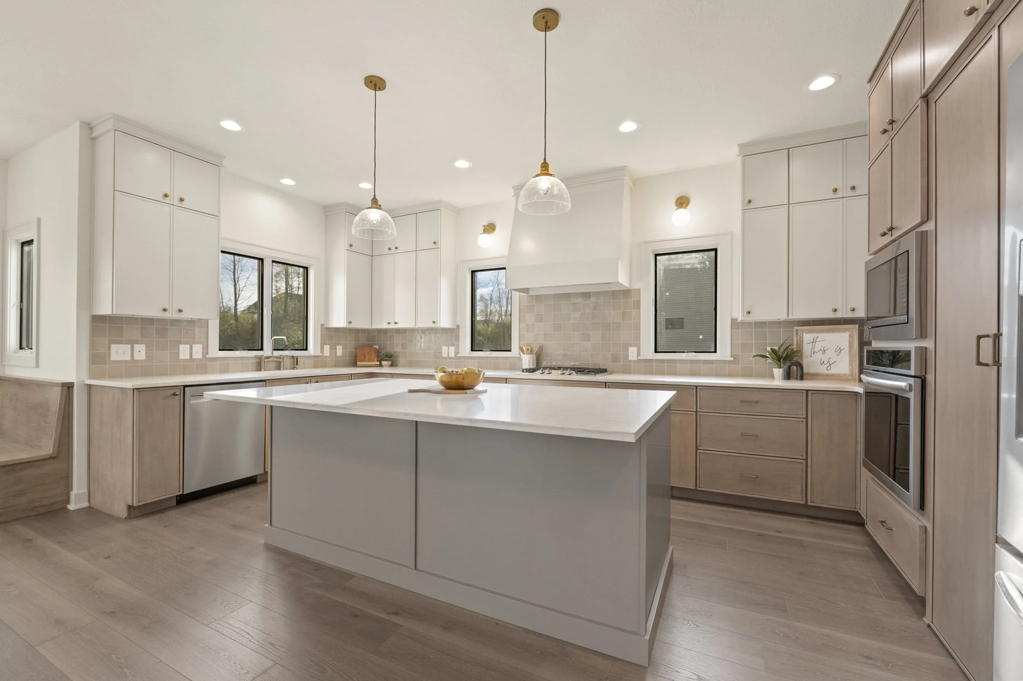 Bright, modern kitchen with white and light wood cabinetry, two pendant lights over a central island, recessed ceiling lights, a window above the sink, and stainless steel appliances.