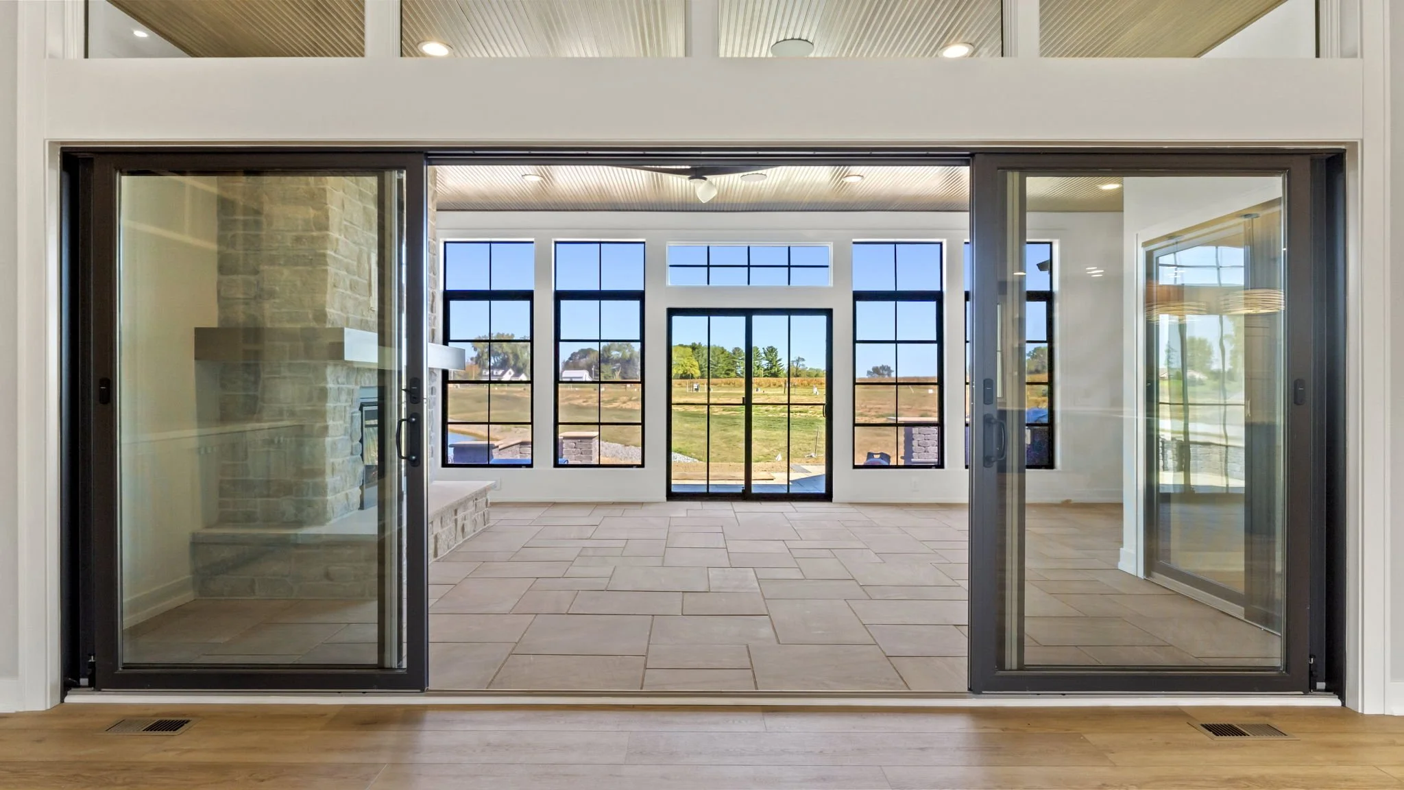 View through sliding glass doors into an interior room with floor-to-ceiling windows, a stone fireplace, and a door leading outside to a yard.